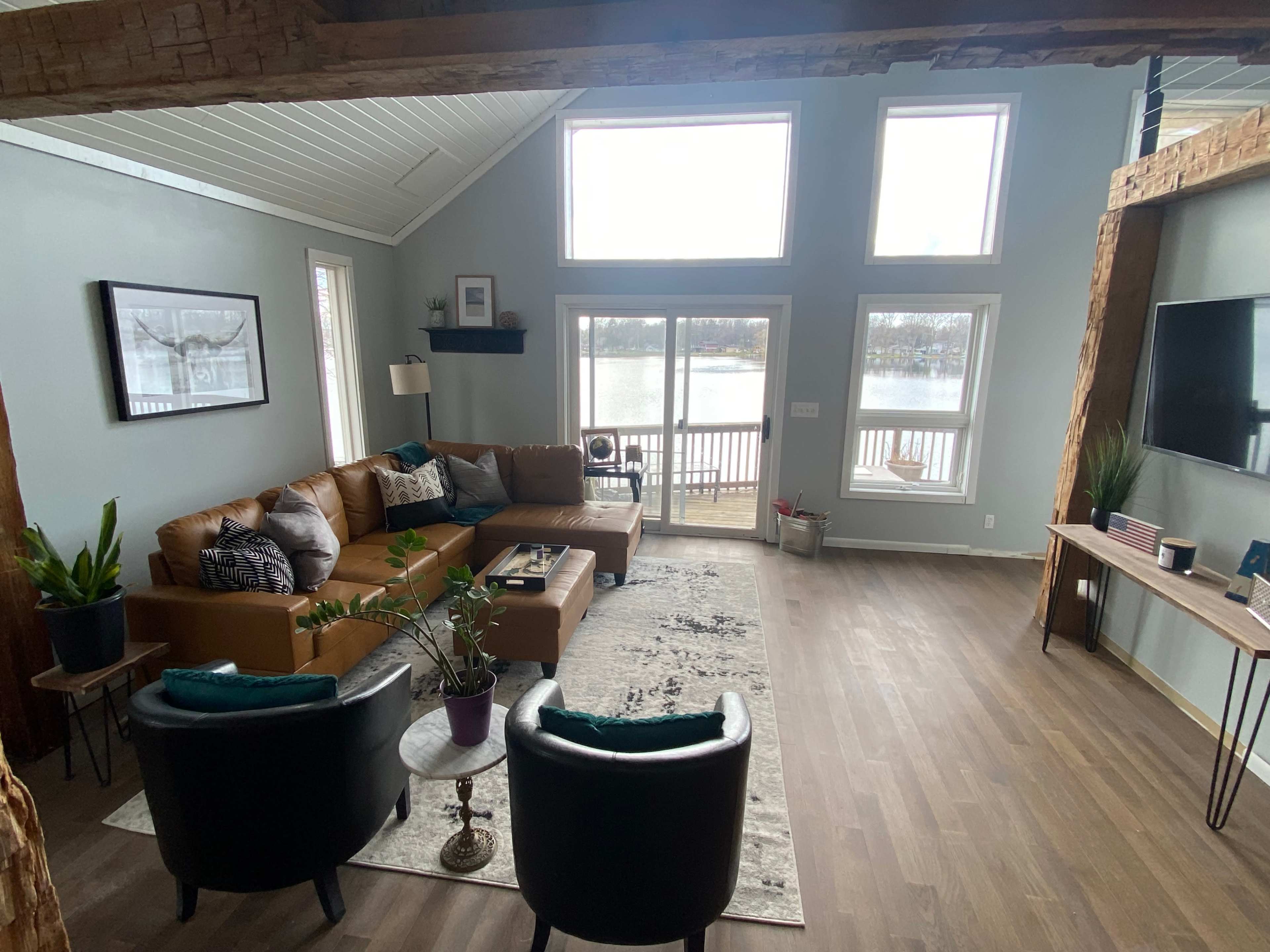 The living room features a brown leather sectional sofa, two black chairs, and large windows that overlook a lake.