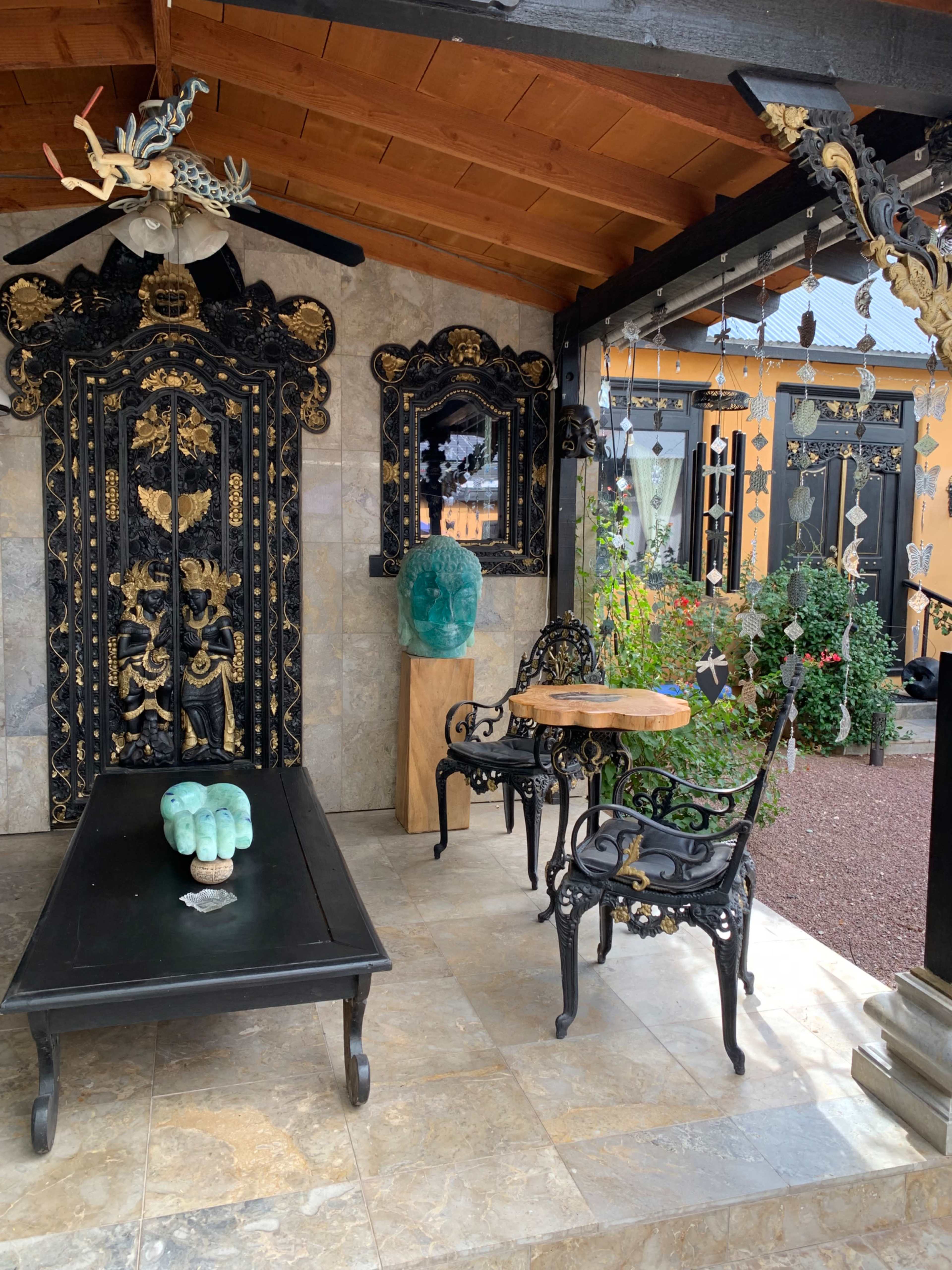 A decorative outdoor space featuring a black intricately carved wooden door, a stone-tiled floor, and wrought iron furniture with a small table and a sculptural green piece on it.