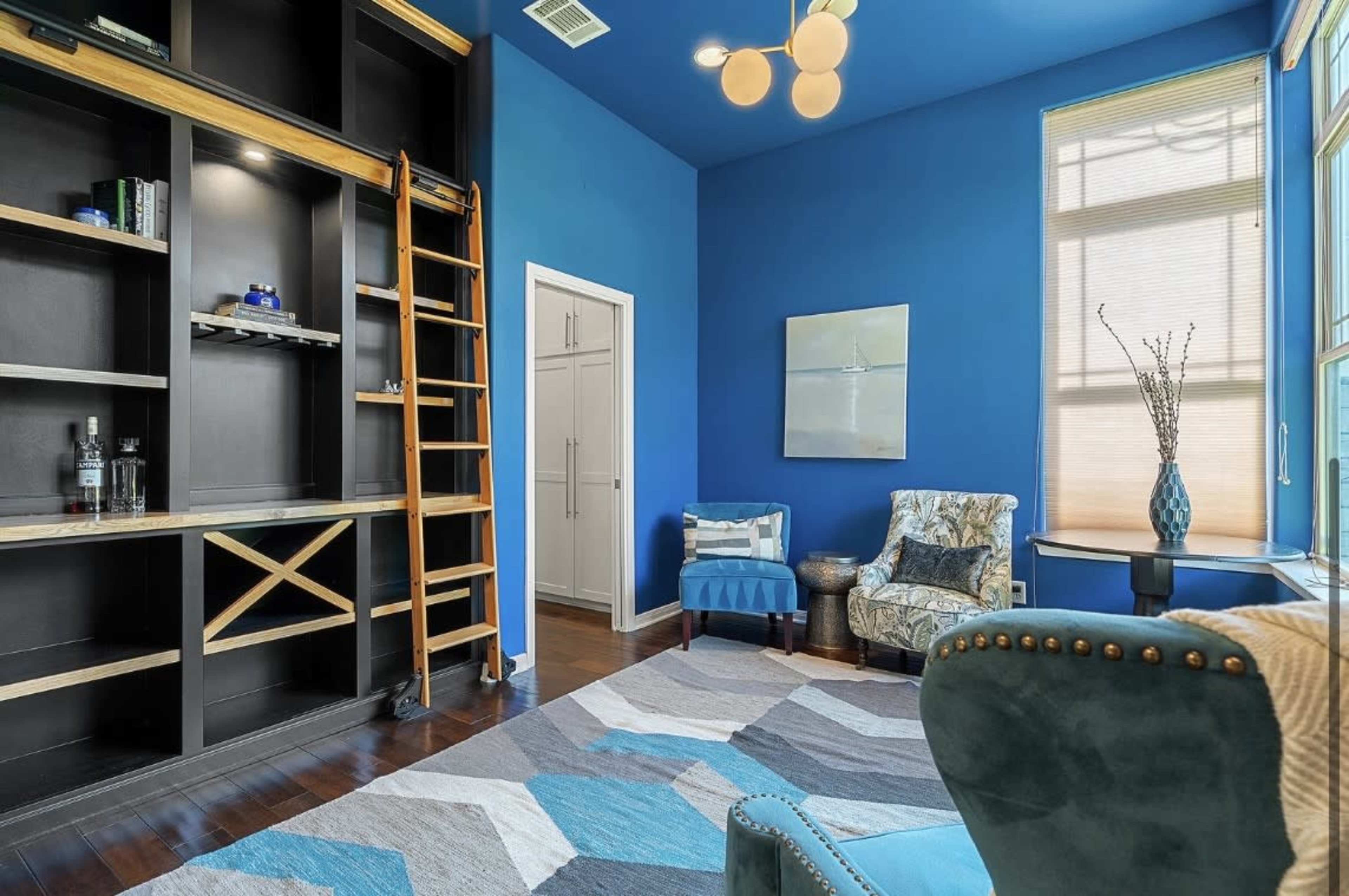 The room features blue walls, a built-in bookshelf with a ladder, and a combination of seating options including a patterned armchair and a blue upholstered chair.