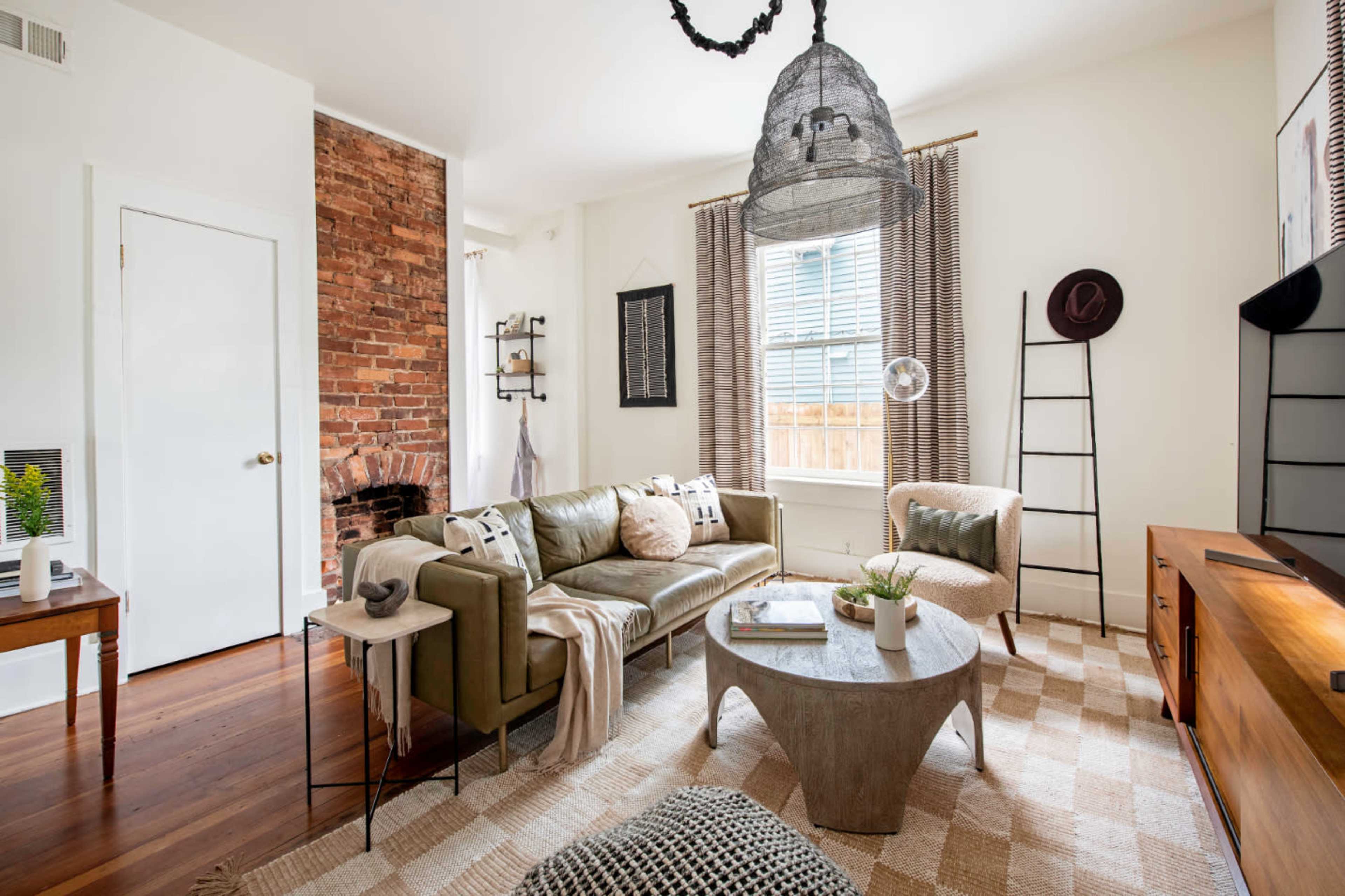 The image shows a modern living room featuring a green sofa, a round coffee table, a mix of brick and white walls, and large windows with curtains.