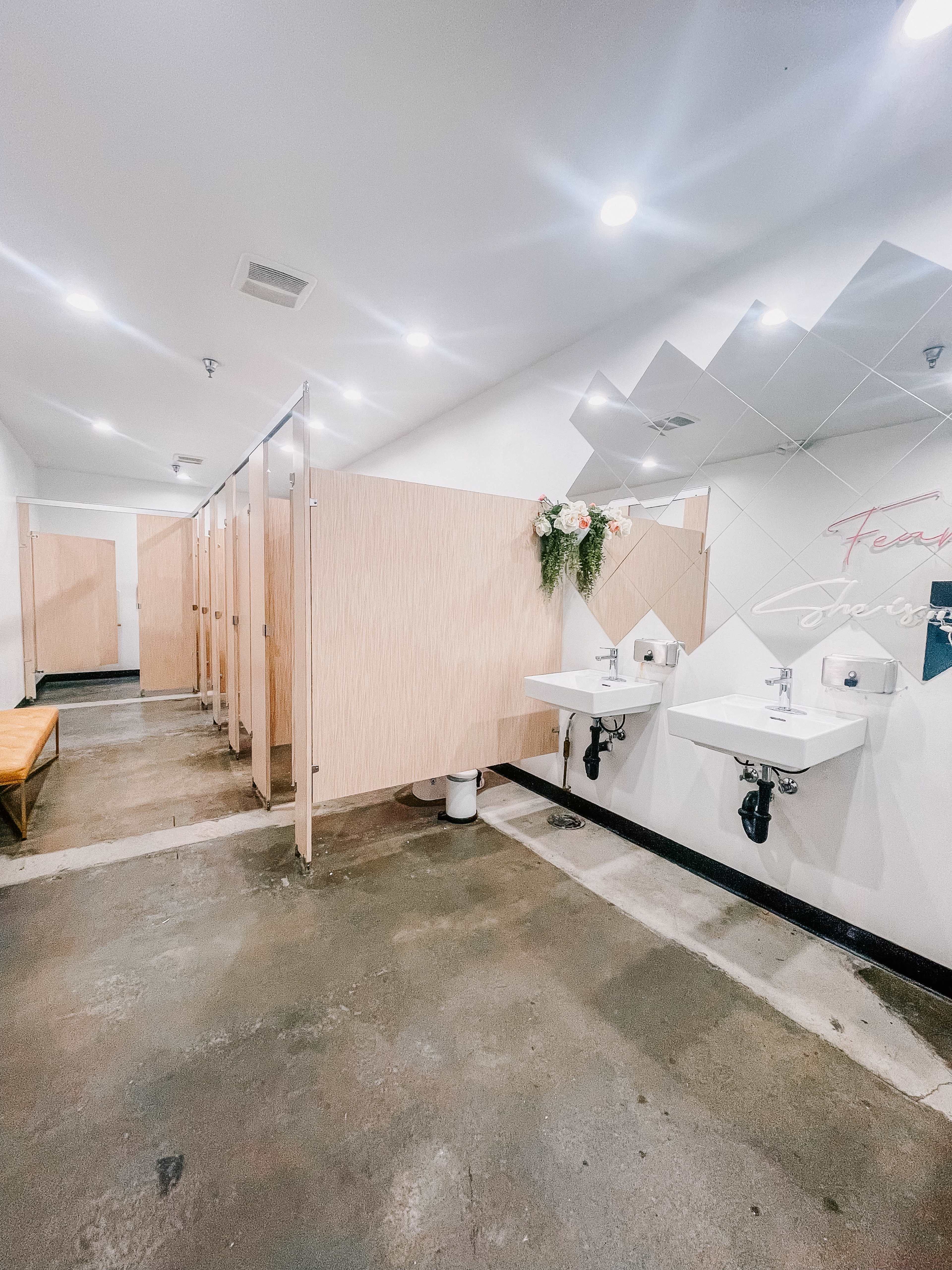A spacious restroom with several partitioned stalls, dual sinks, and minimal decor, featuring a plant arrangement above a mirror.