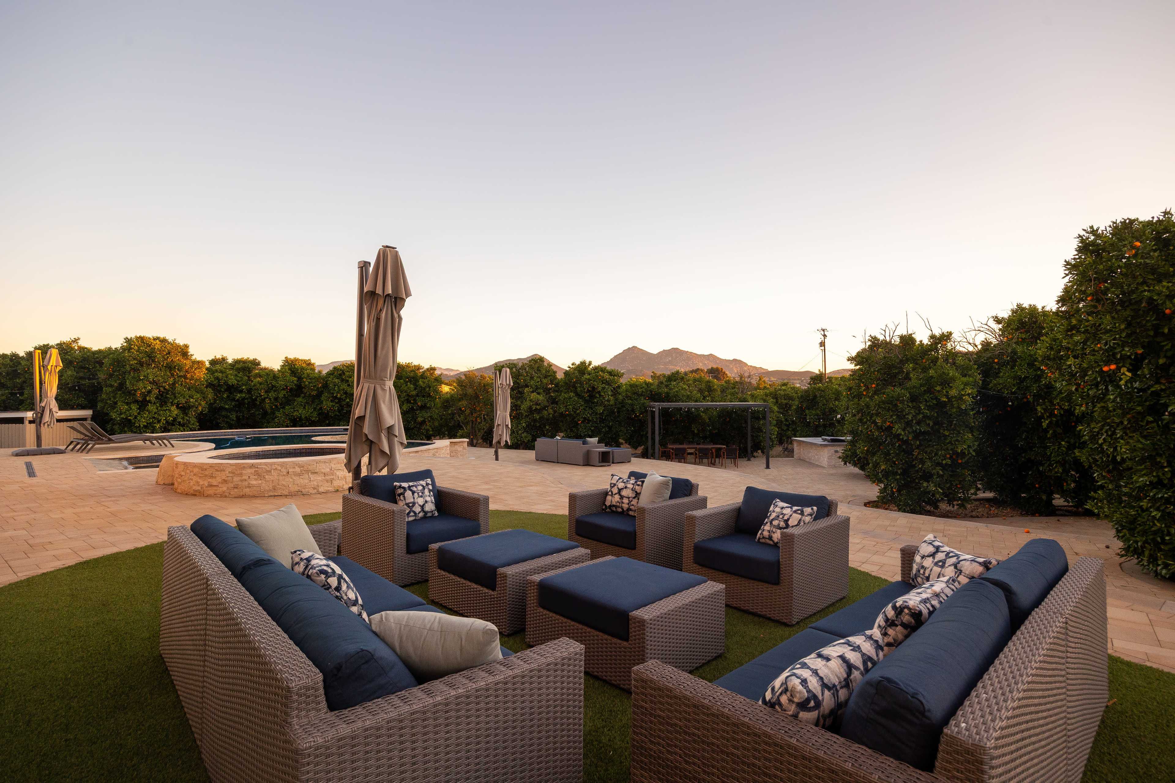A seating area with woven furniture and cushions is set on a patio beside a pool, framed by greenery and mountains in the background.