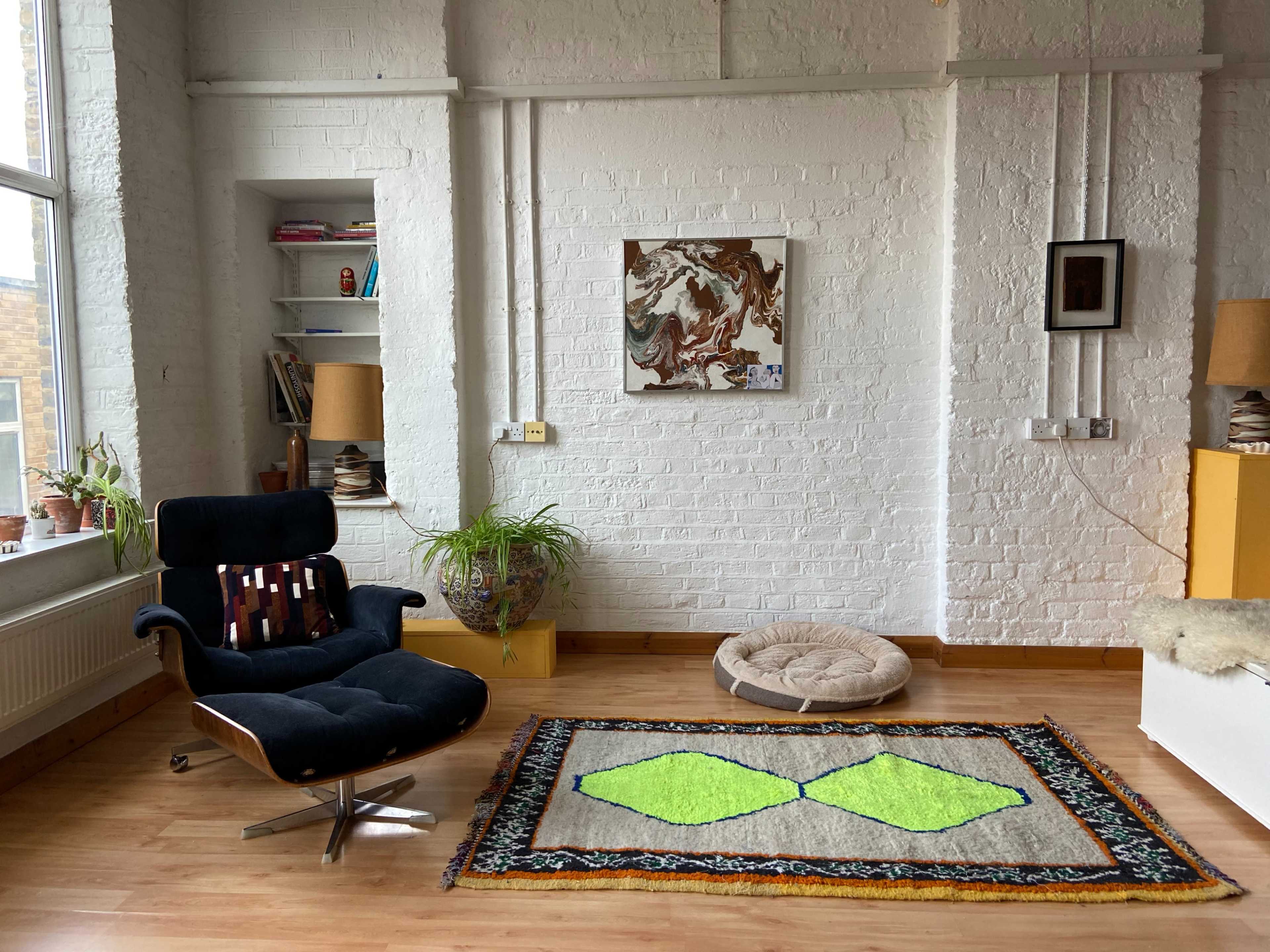 The room features a modern black chair, a colorful area rug with a geometric design, a round pet bed, and shelves with books and plants against a white brick wall.