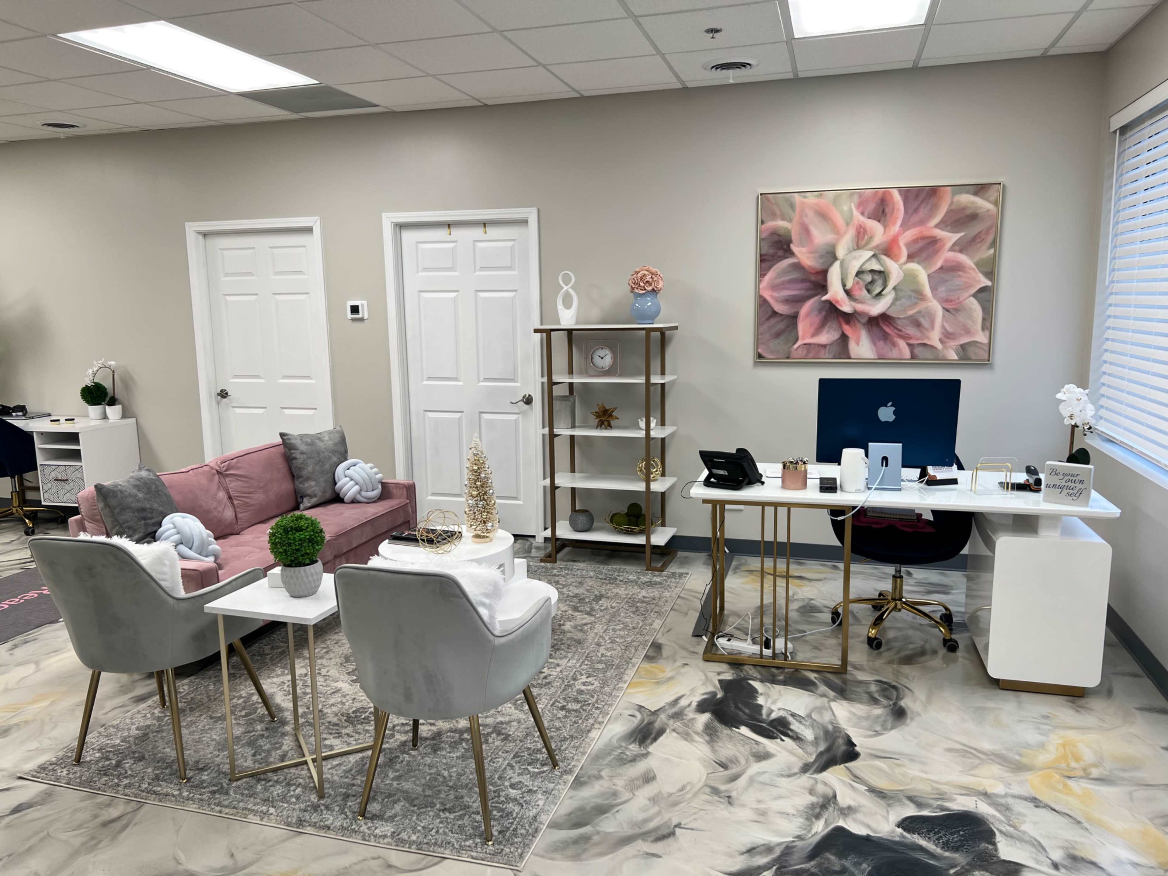 A modern office space featuring a seating area with a pink sofa and two chairs, a decorative bookshelf, and a desk with a computer against a light-colored wall.