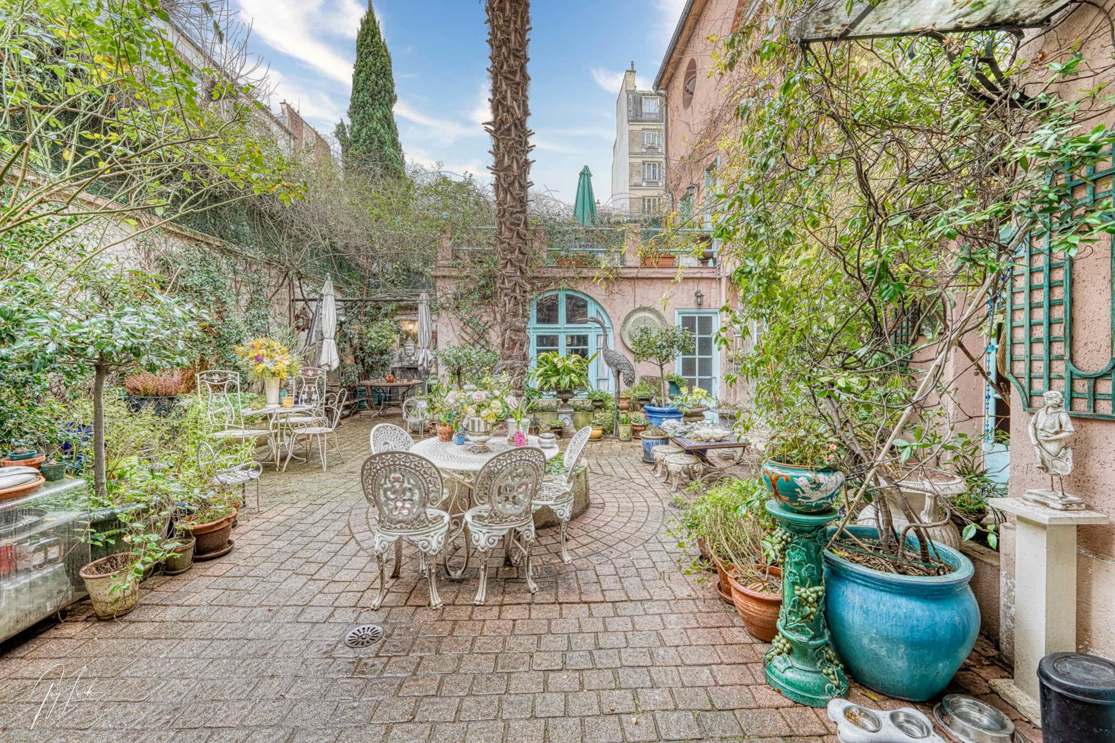 A lush courtyard garden with various potted plants, stone pathways, and a table set with chairs under a palm tree.