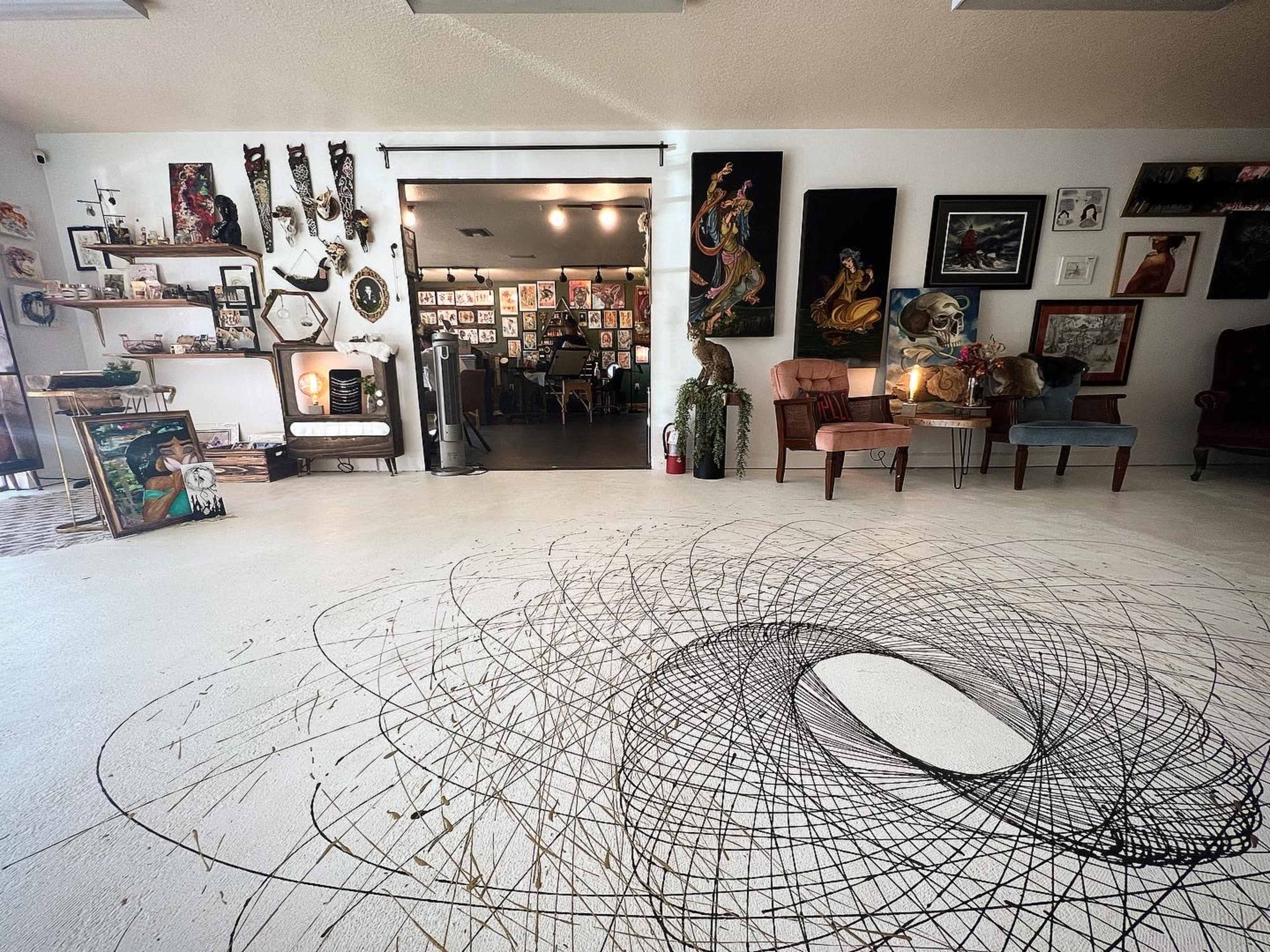 An art studio with a spiraling black pattern on the white floor, surrounded by various framed artworks and decorative items on the walls.