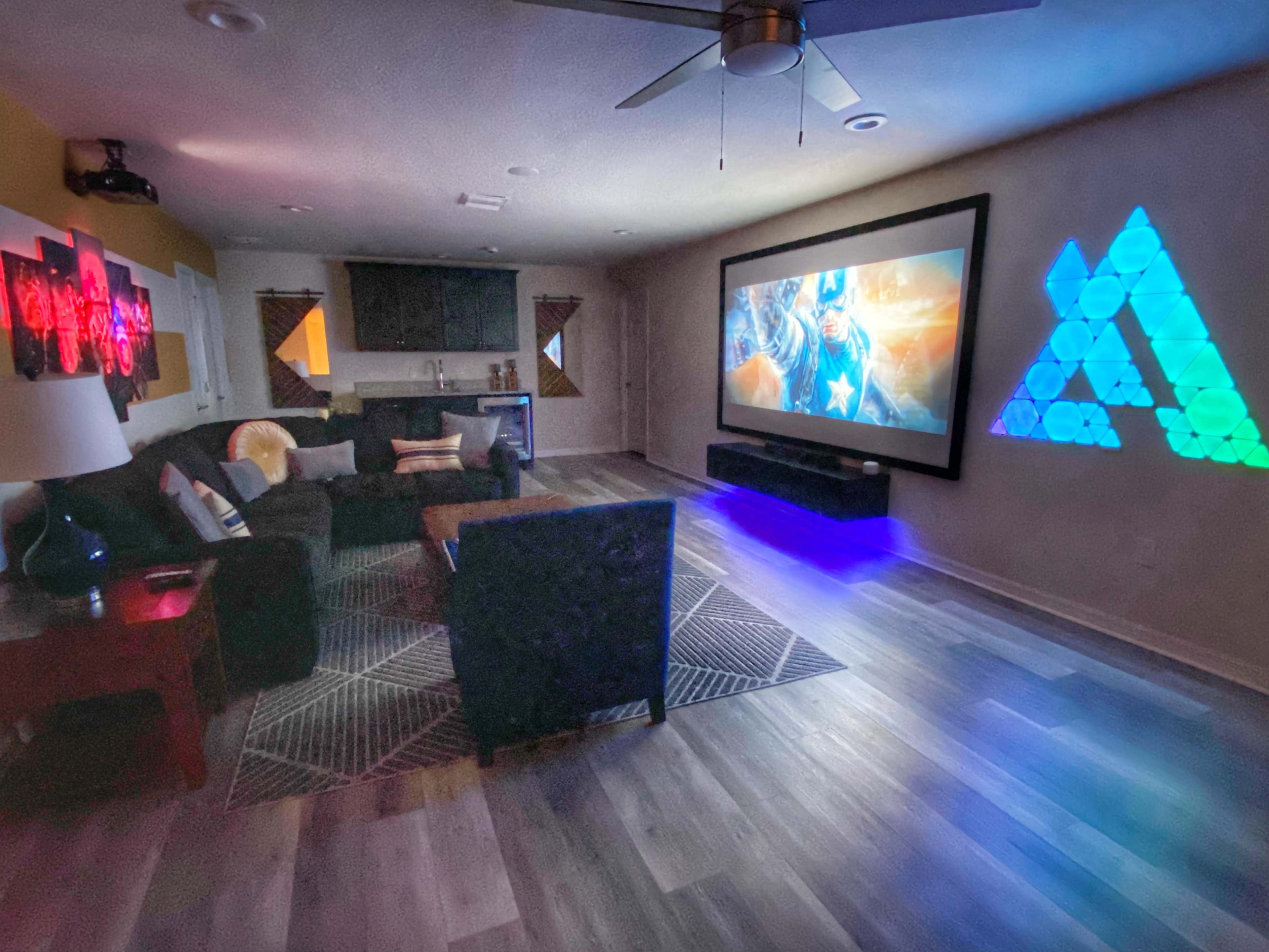 The image shows a modern living room with a large projection screen displaying a superhero theme, accompanied by colorful lighting accents and a cozy seating area.