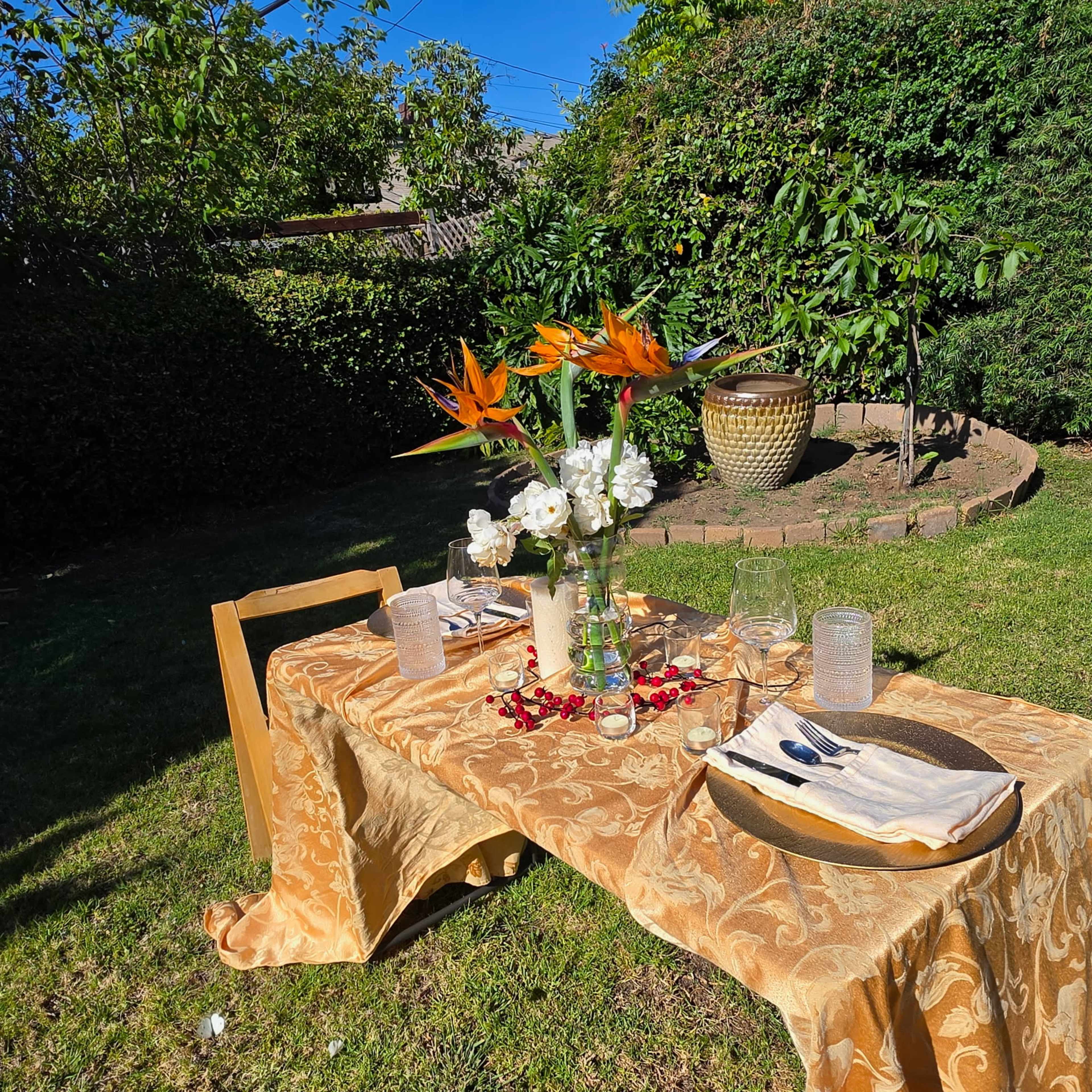 A neatly set outdoor table with a decorative tablecloth, glassware, and a centerpiece of flowers is positioned in a green garden.
