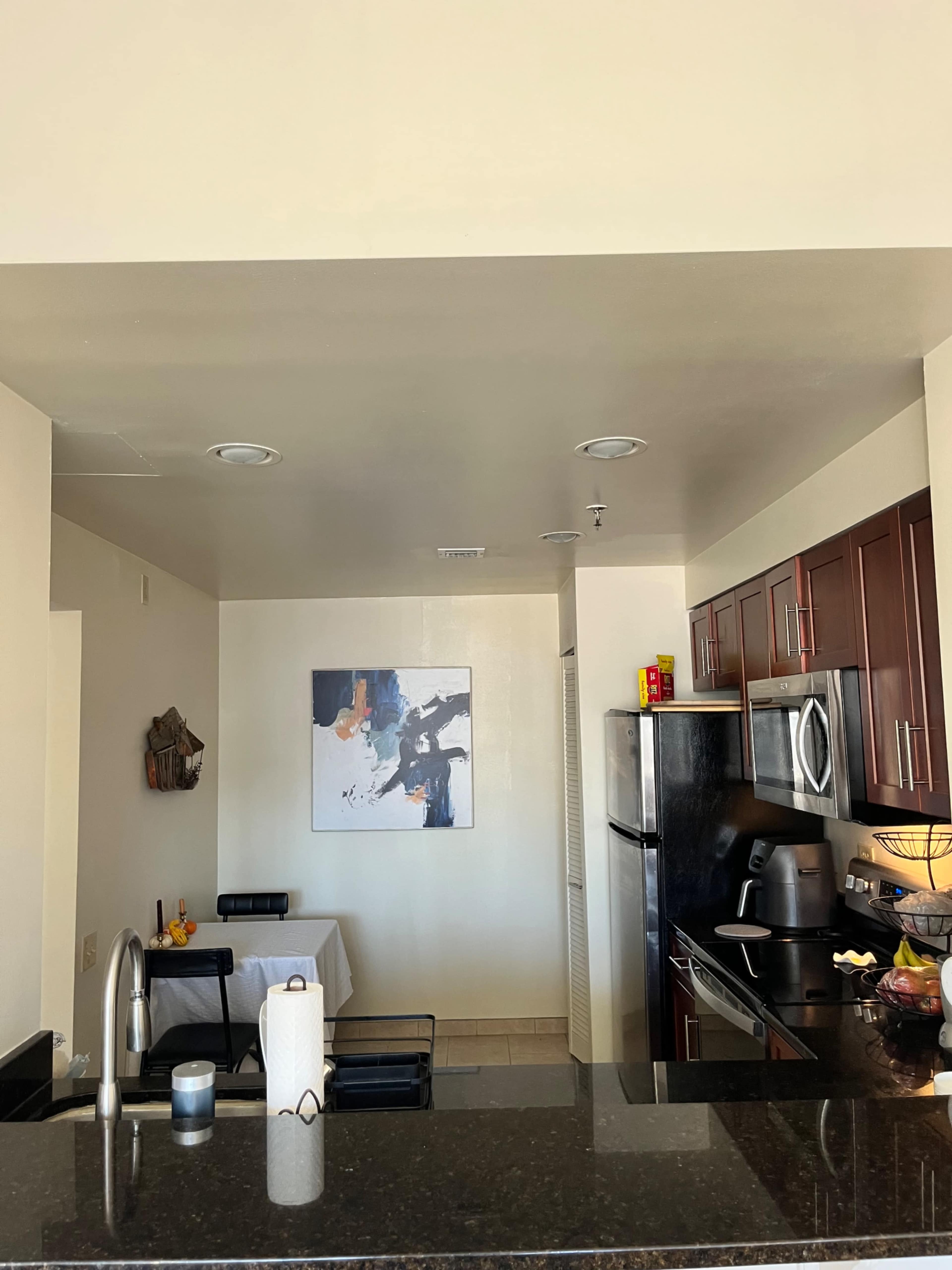 The image shows a modern kitchen with dark wood cabinets, a stainless steel refrigerator, and an adjacent dining area with a small table and chairs.