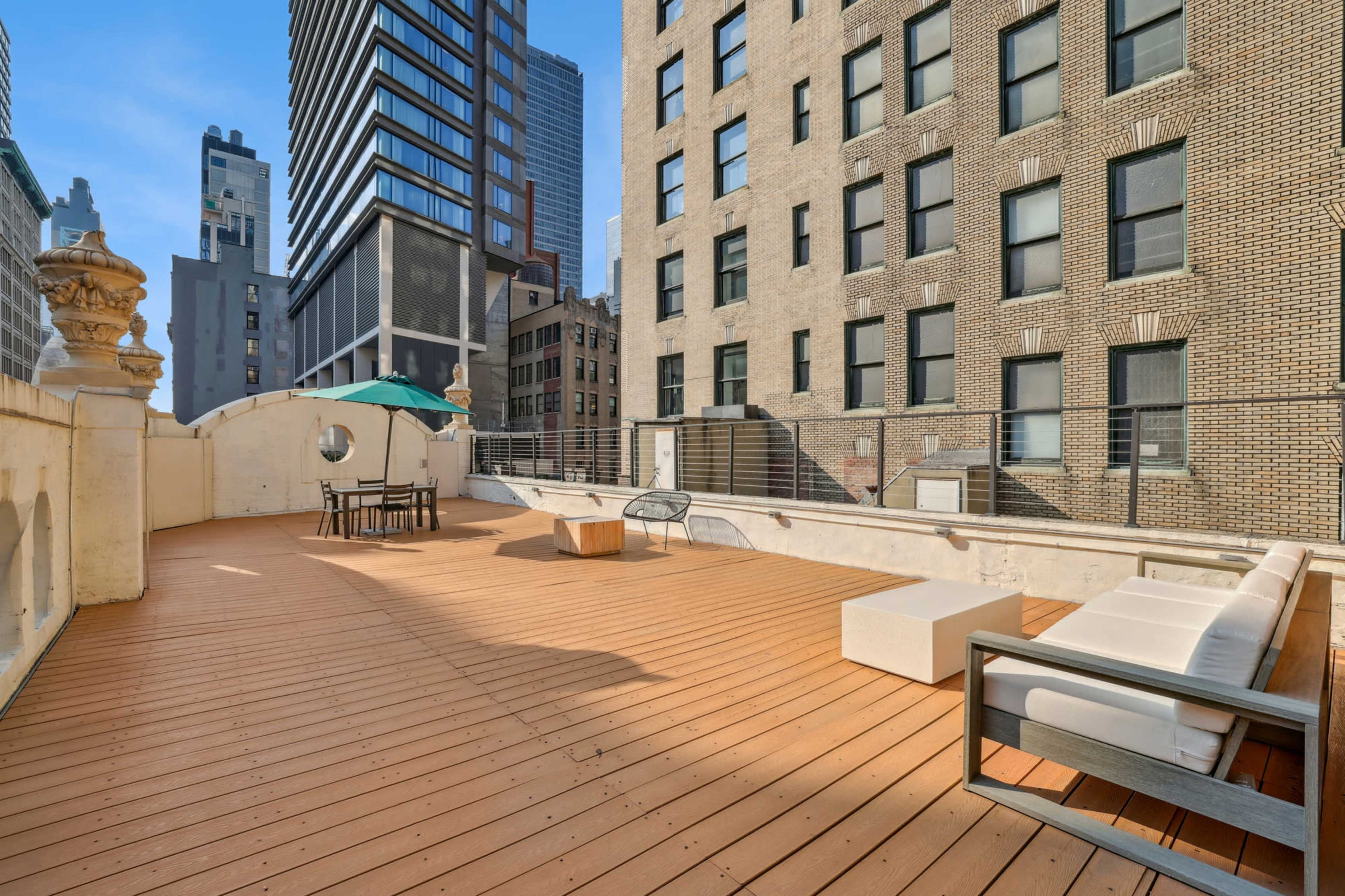 The image shows a spacious rooftop terrace featuring wooden decking, a seating area, and views of surrounding buildings in an urban setting.