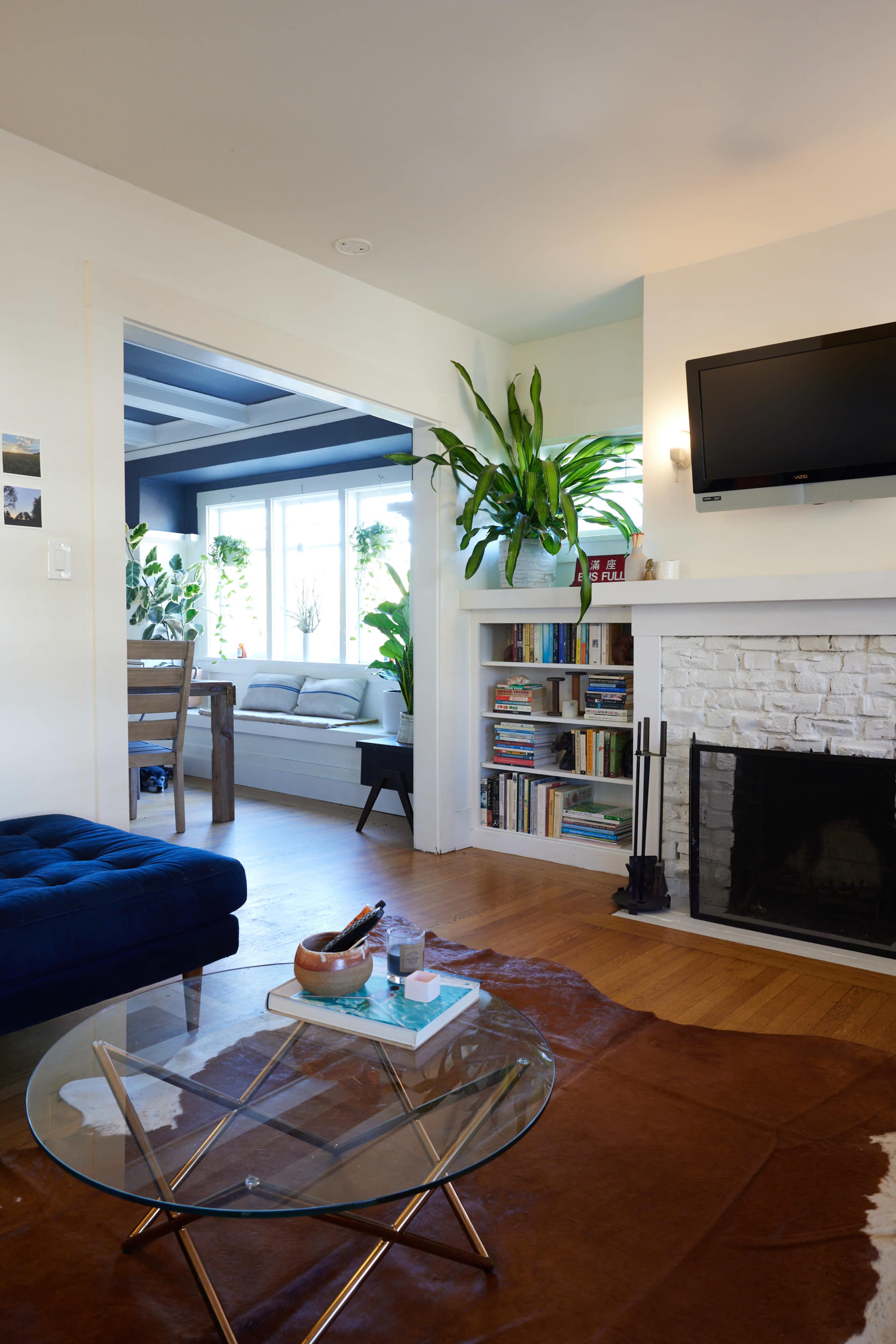 A bright living room featuring a blue velvet ottoman, a glass coffee table on a patterned rug, and a fireplace with a television mounted above it.