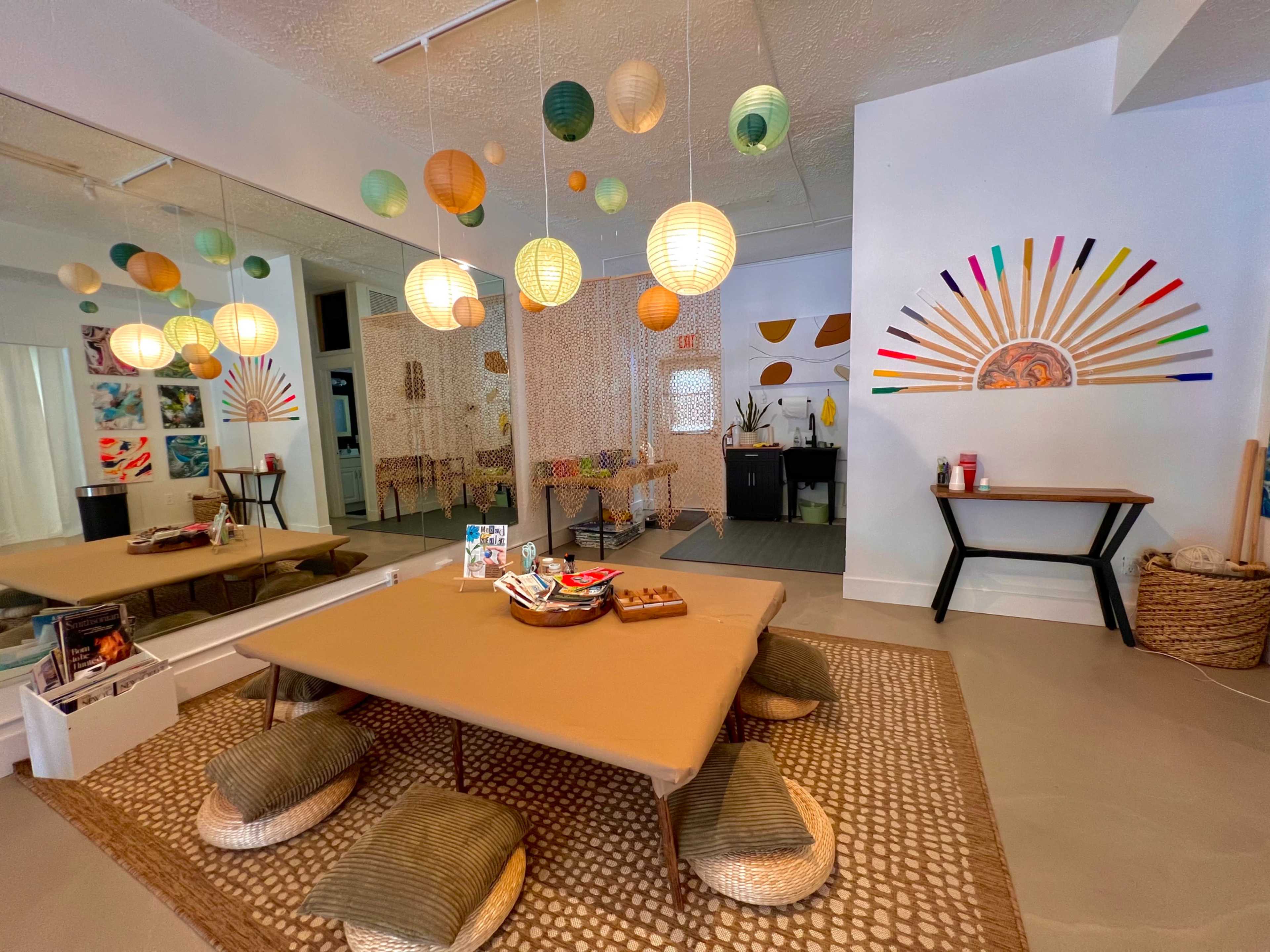 A spacious room featuring a low table surrounded by floor cushions, with decorative paper lanterns hanging from the ceiling and a large mural on one wall.