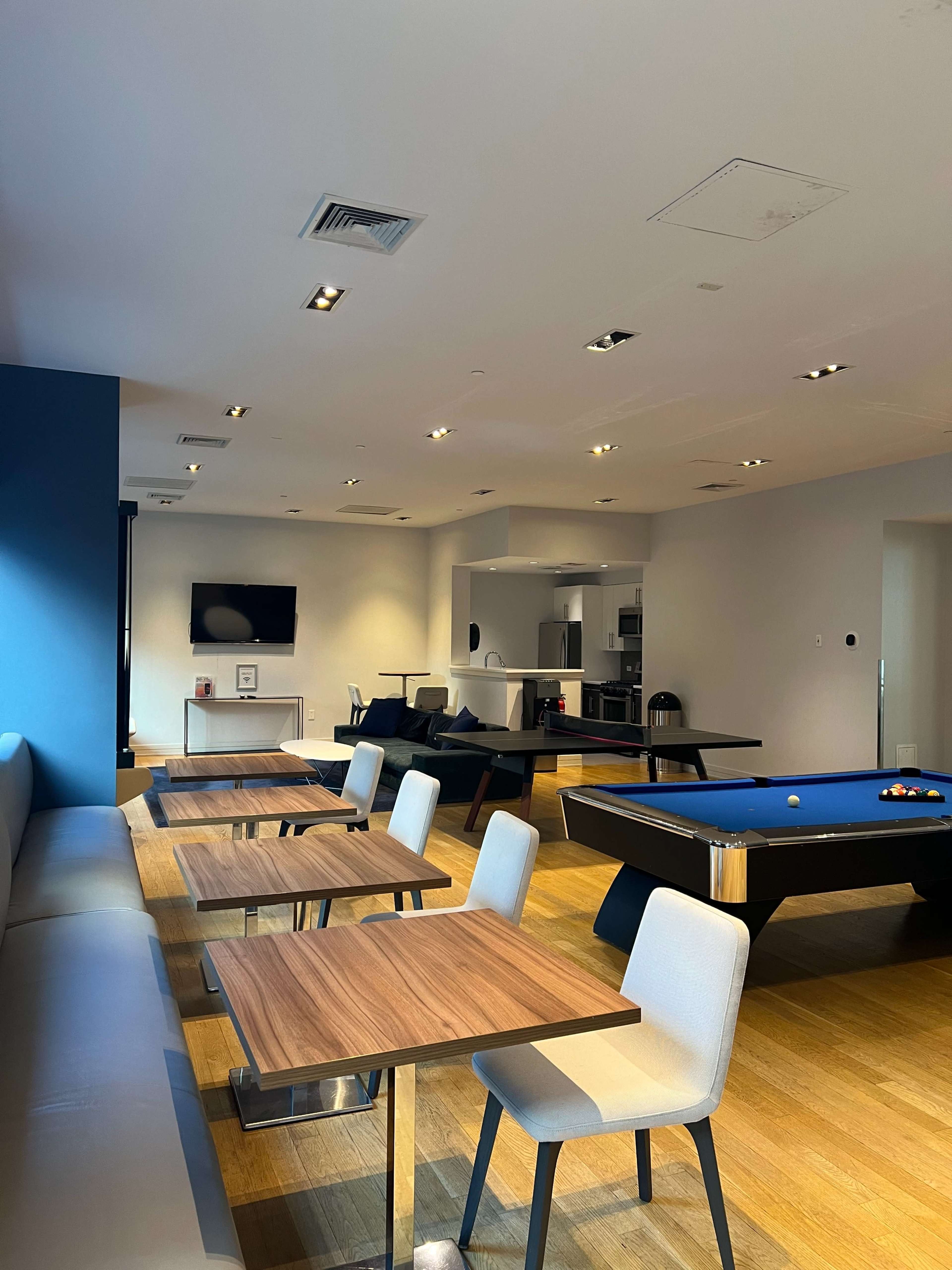 A communal lounge area featuring several wooden tables and chairs, a billiards table, and a kitchenette in the background.