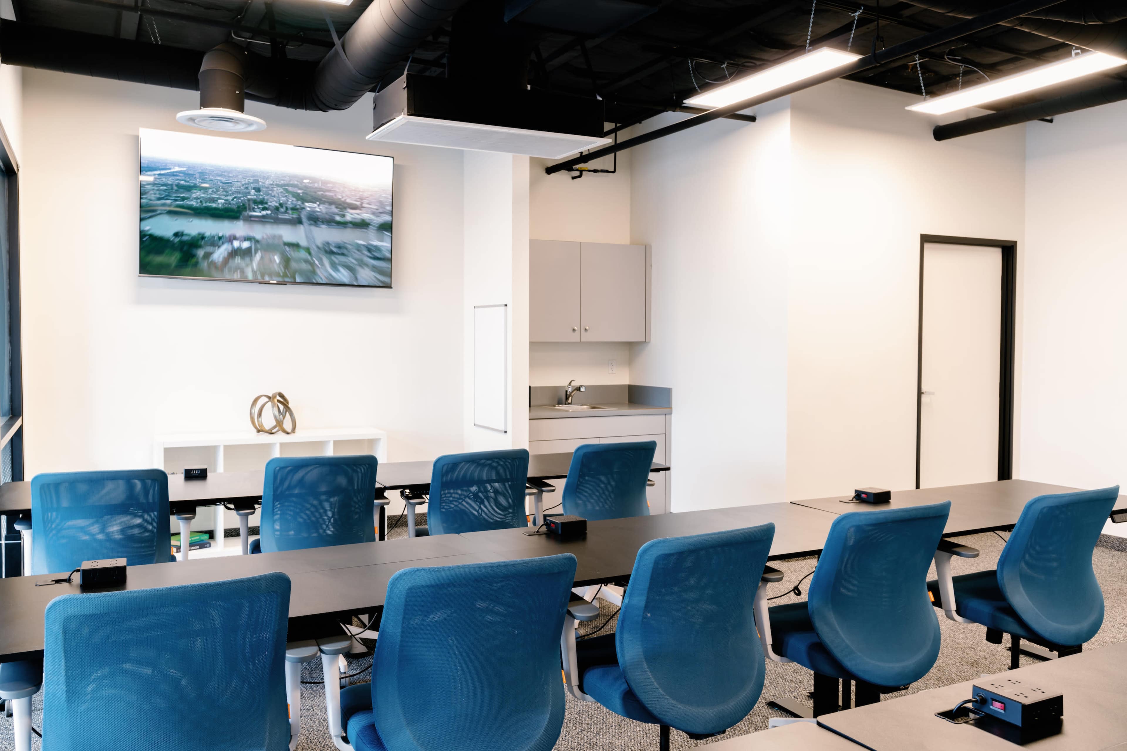 A modern conference room features a long table with blue chairs, a wall-mounted screen displaying a video, and a kitchenette area in the background.