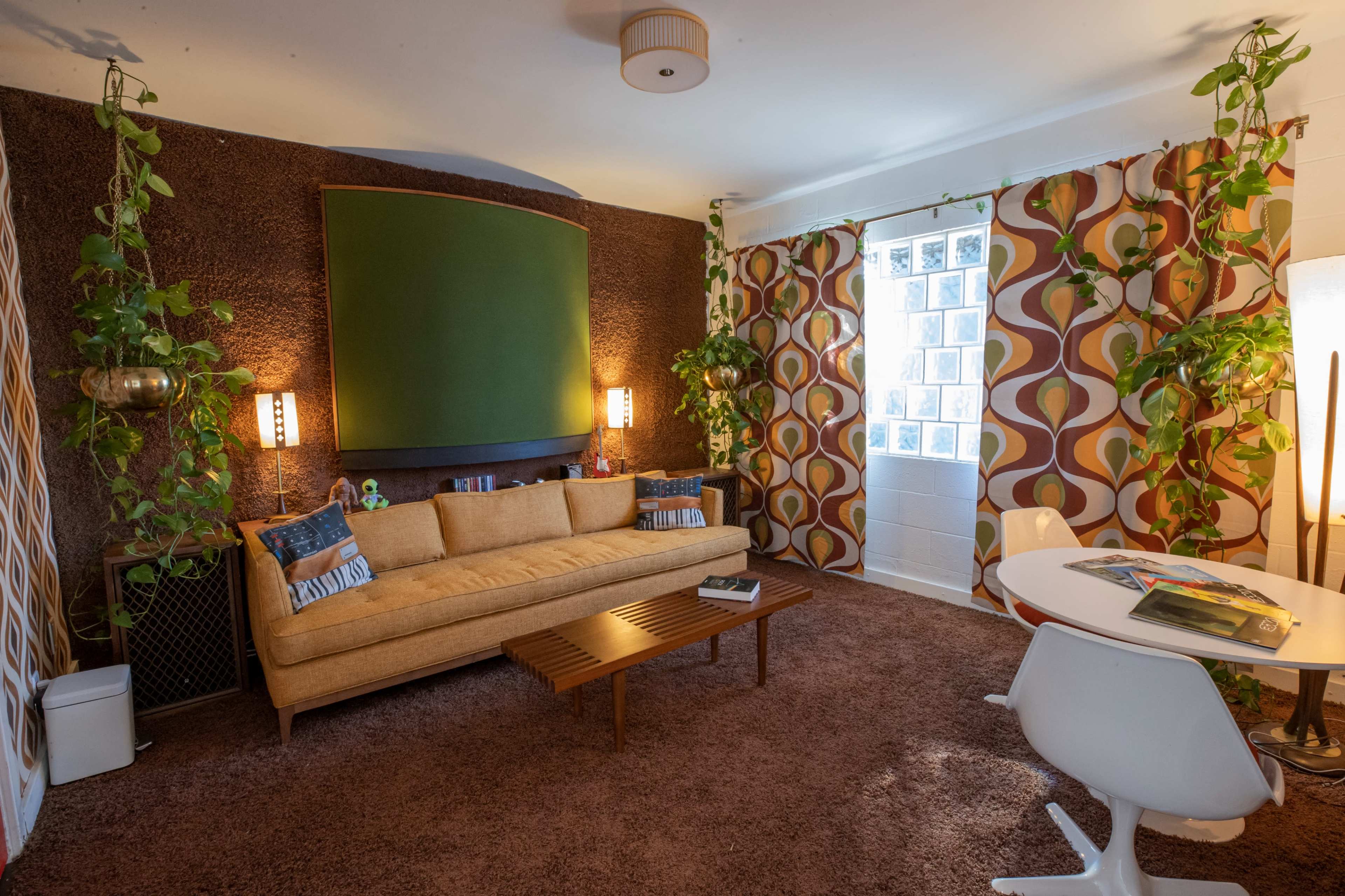 A living room with a yellow couch, a wooden coffee table, patterned curtains, and various green plants.
