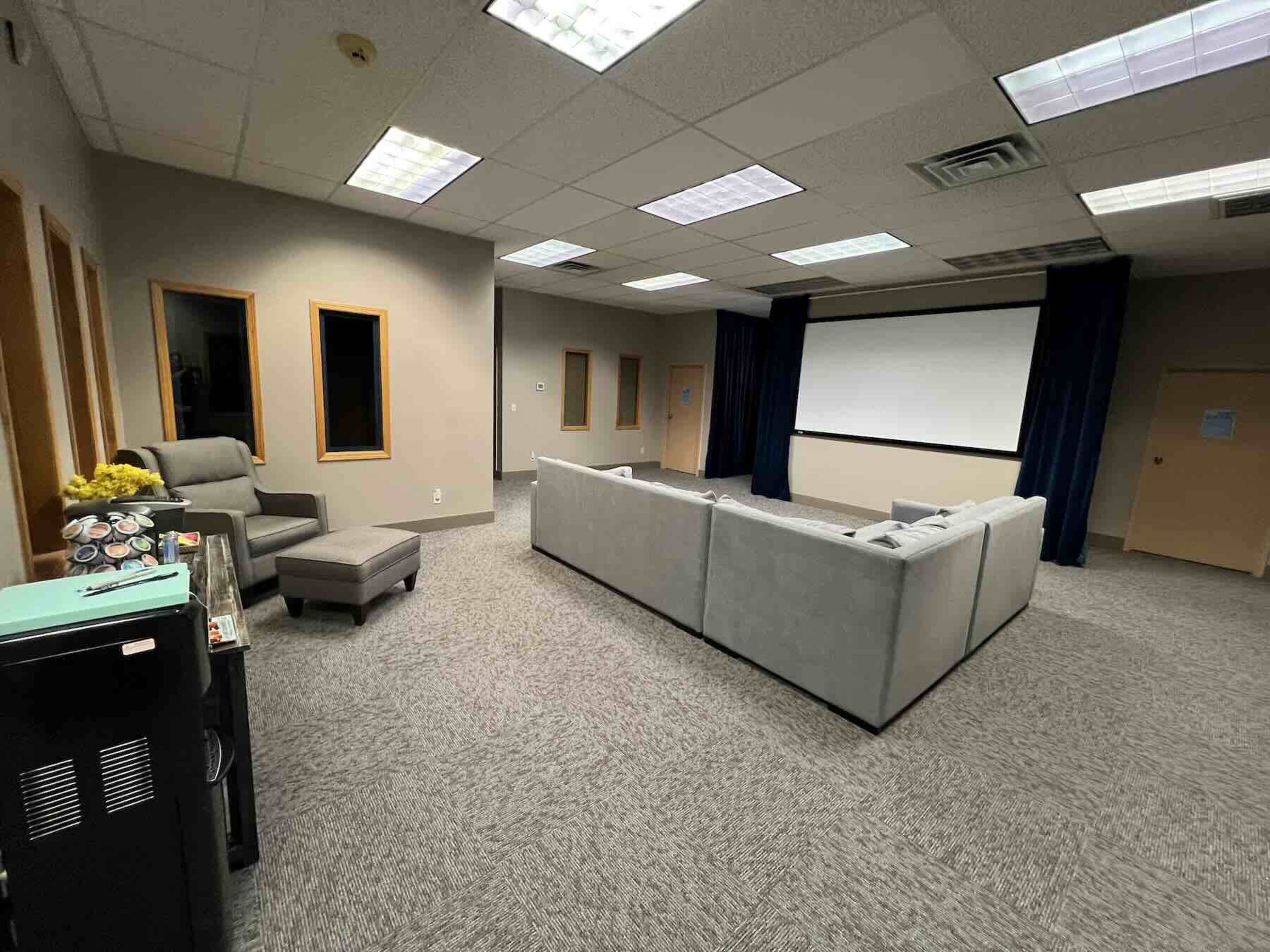 The image shows a conference room with a gray sectional sofa facing a large screen, accompanied by a chair and two windows.