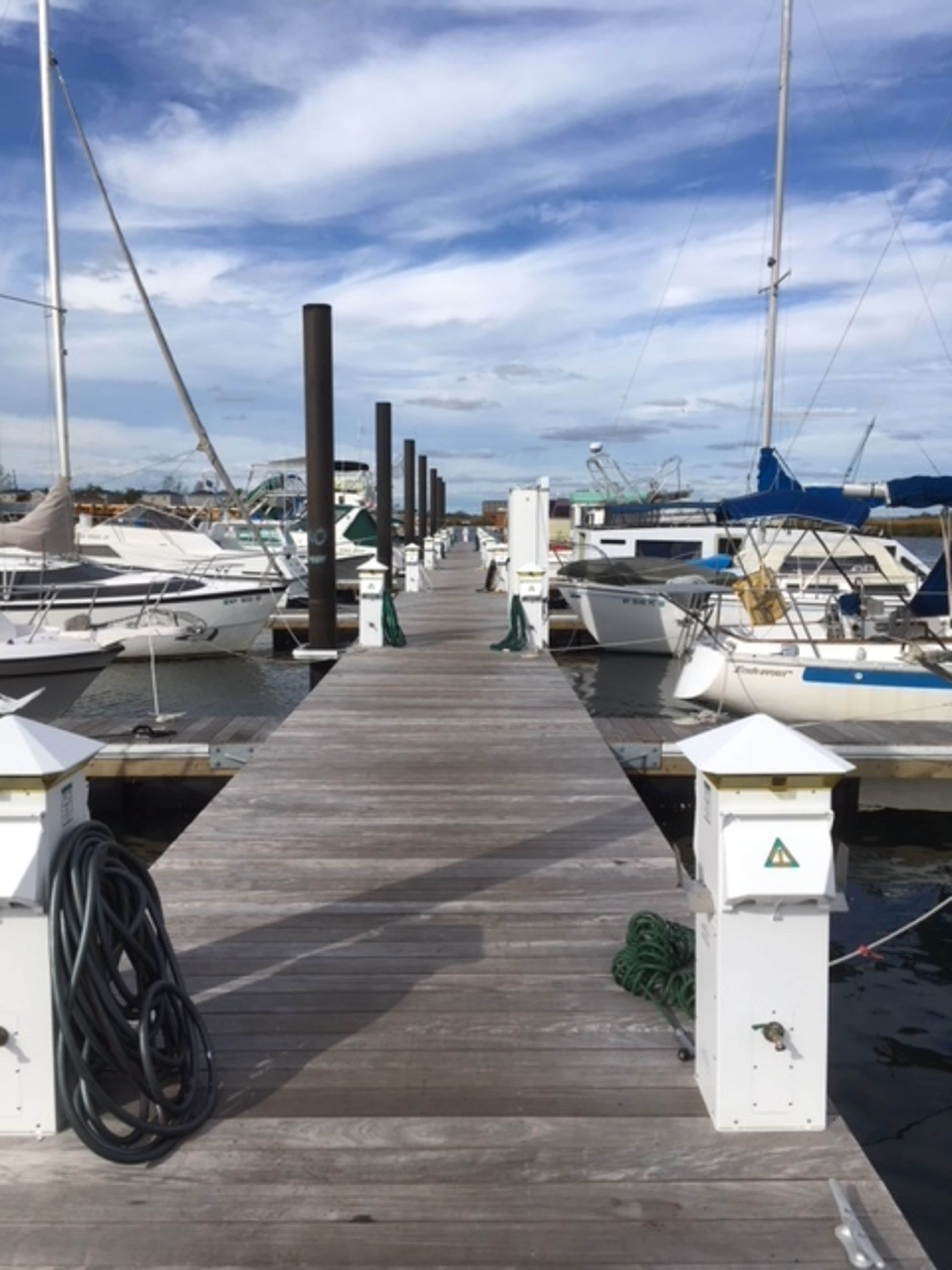 A wooden dock extends between rows of moored boats, with features like tall posts and clear blue skies overhead.