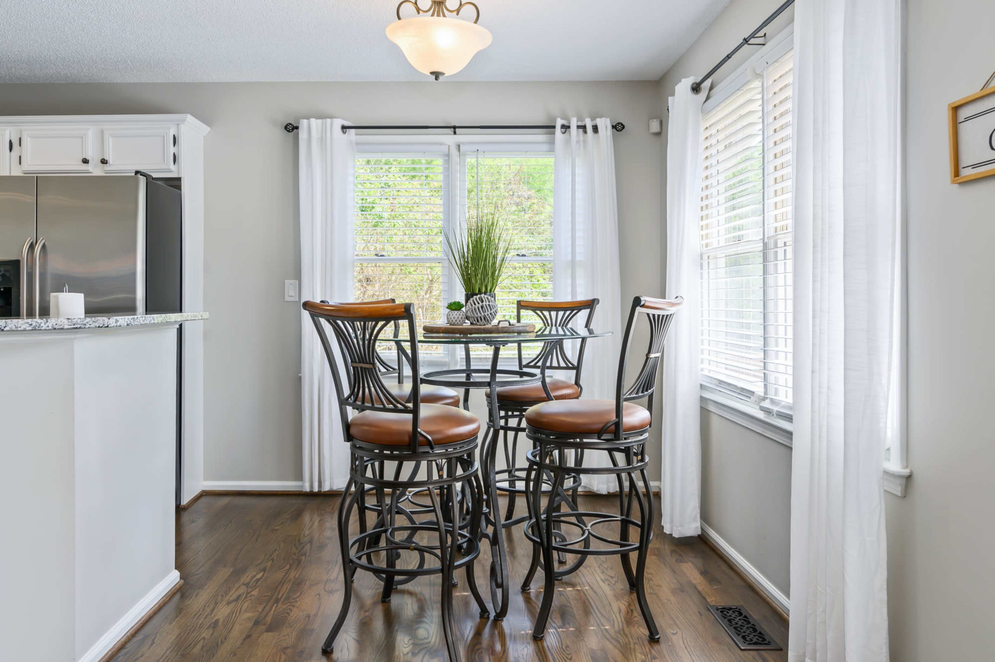 A breakfast nook features a round glass table surrounded by four metal bar stools, with large windows letting in natural light and potted plants as decor.