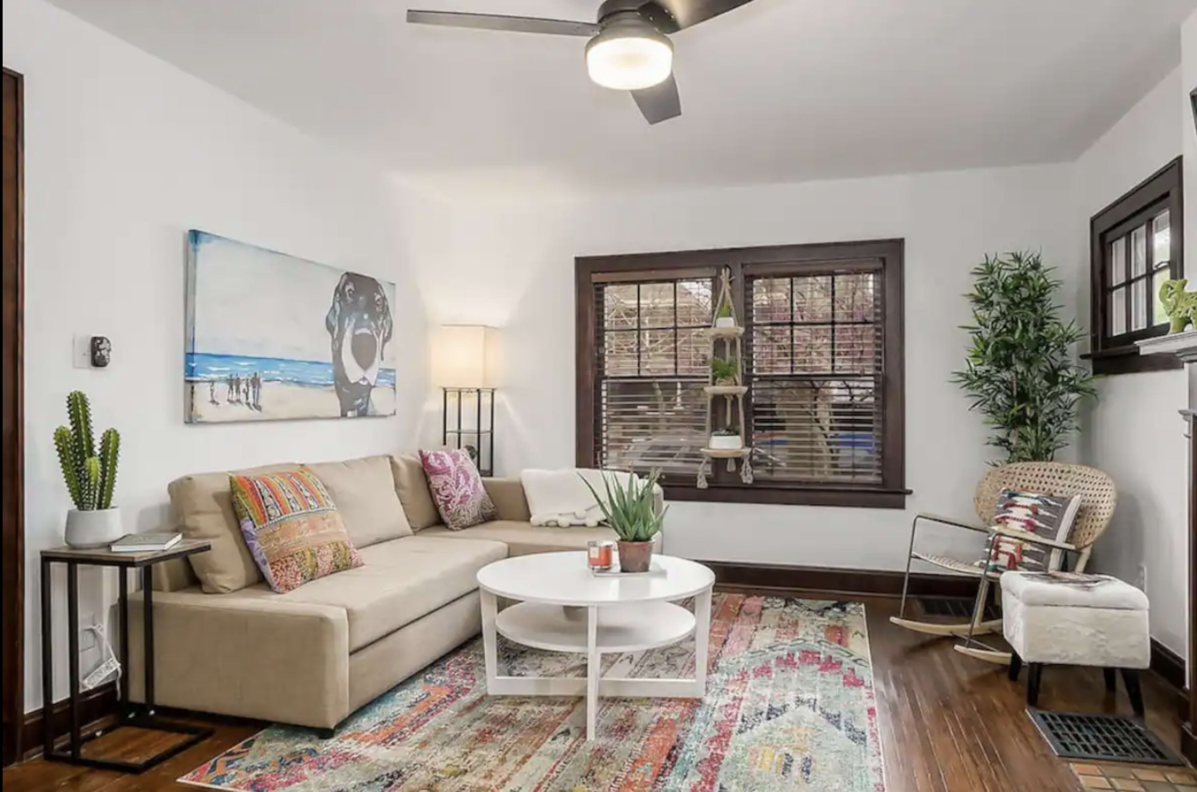 A living room featuring a beige sectional sofa, a round white coffee table, a patterned area rug, and large windows with blinds, complemented by a small indoor plant and decorative items.