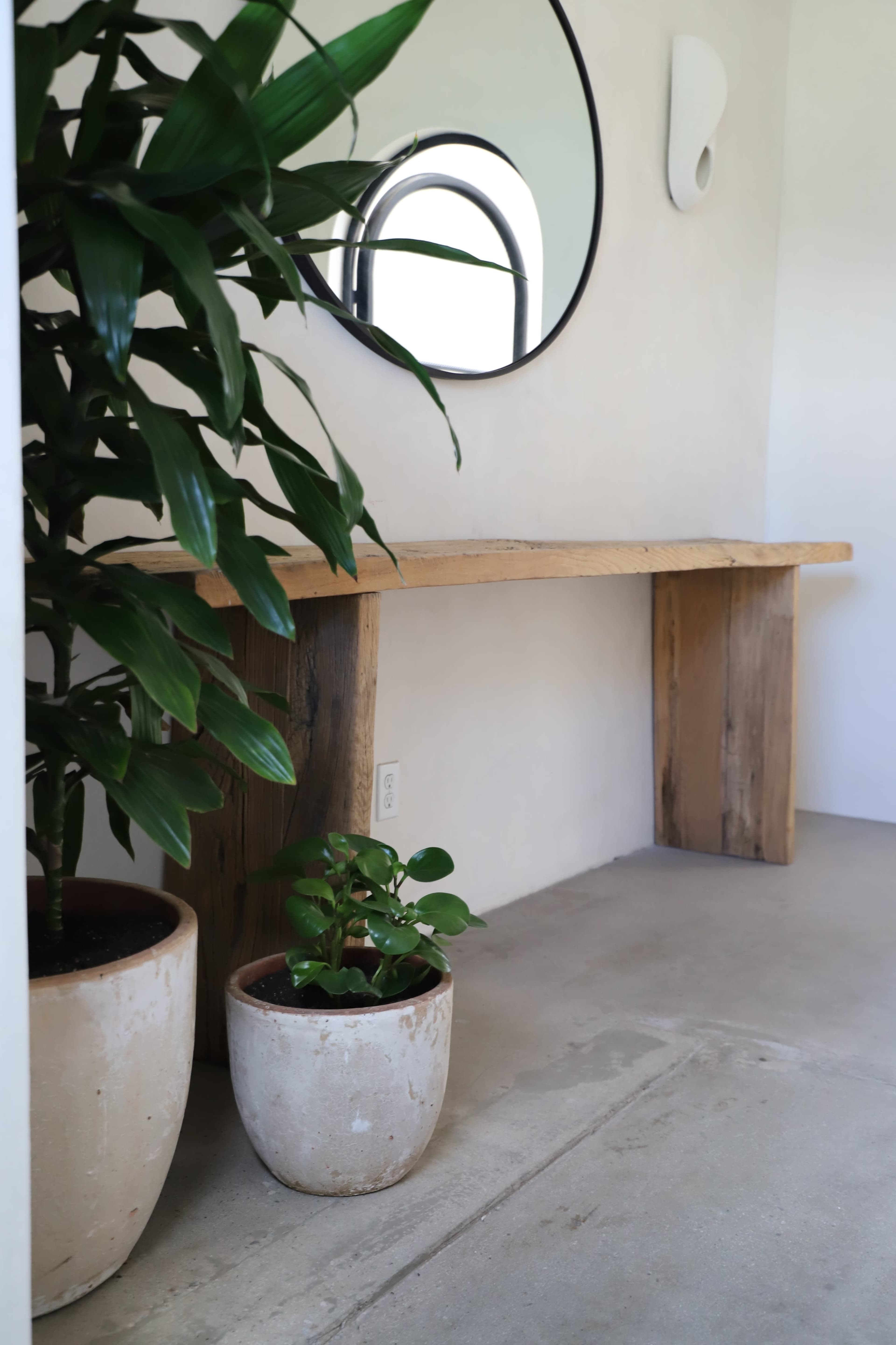 A wooden table with a round mirror above it is situated next to two potted plants in a minimalist interior space.