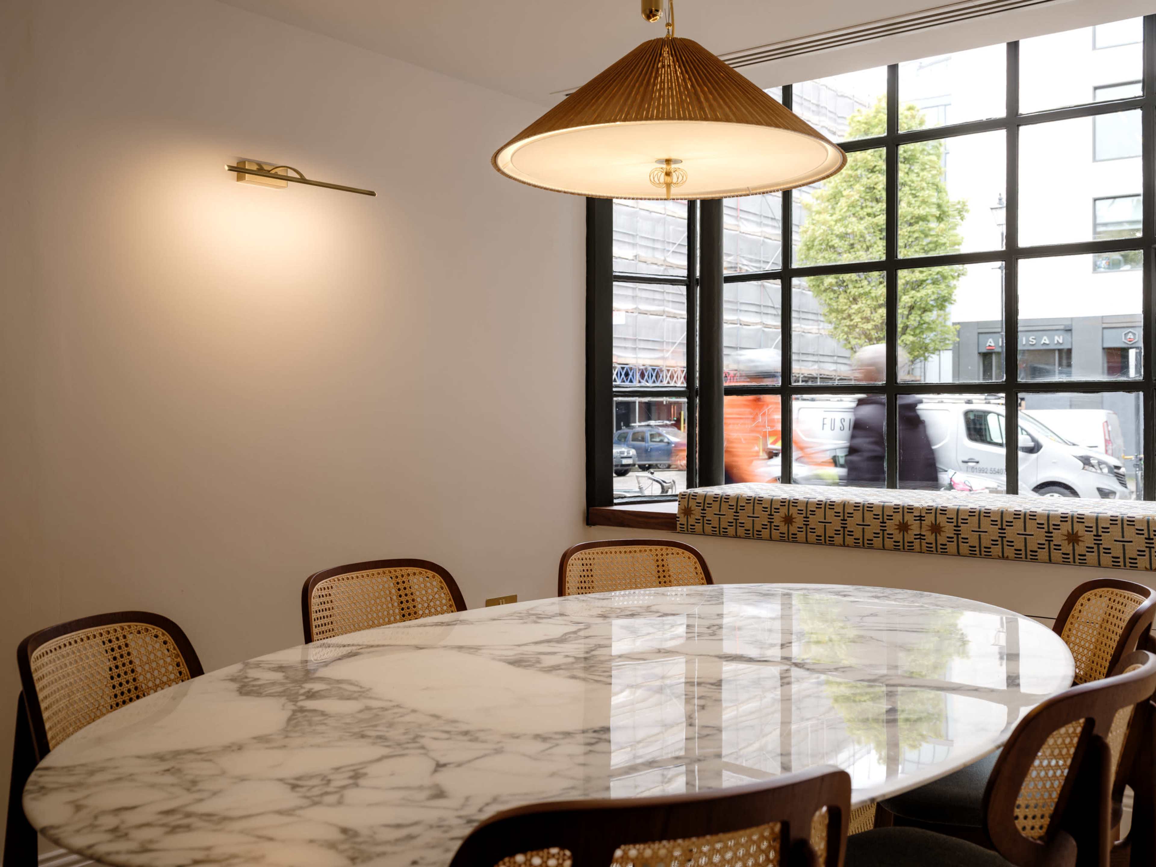 A round marble table surrounded by cane-back chairs is illuminated by a pendant light in a room with a large window displaying a street view.