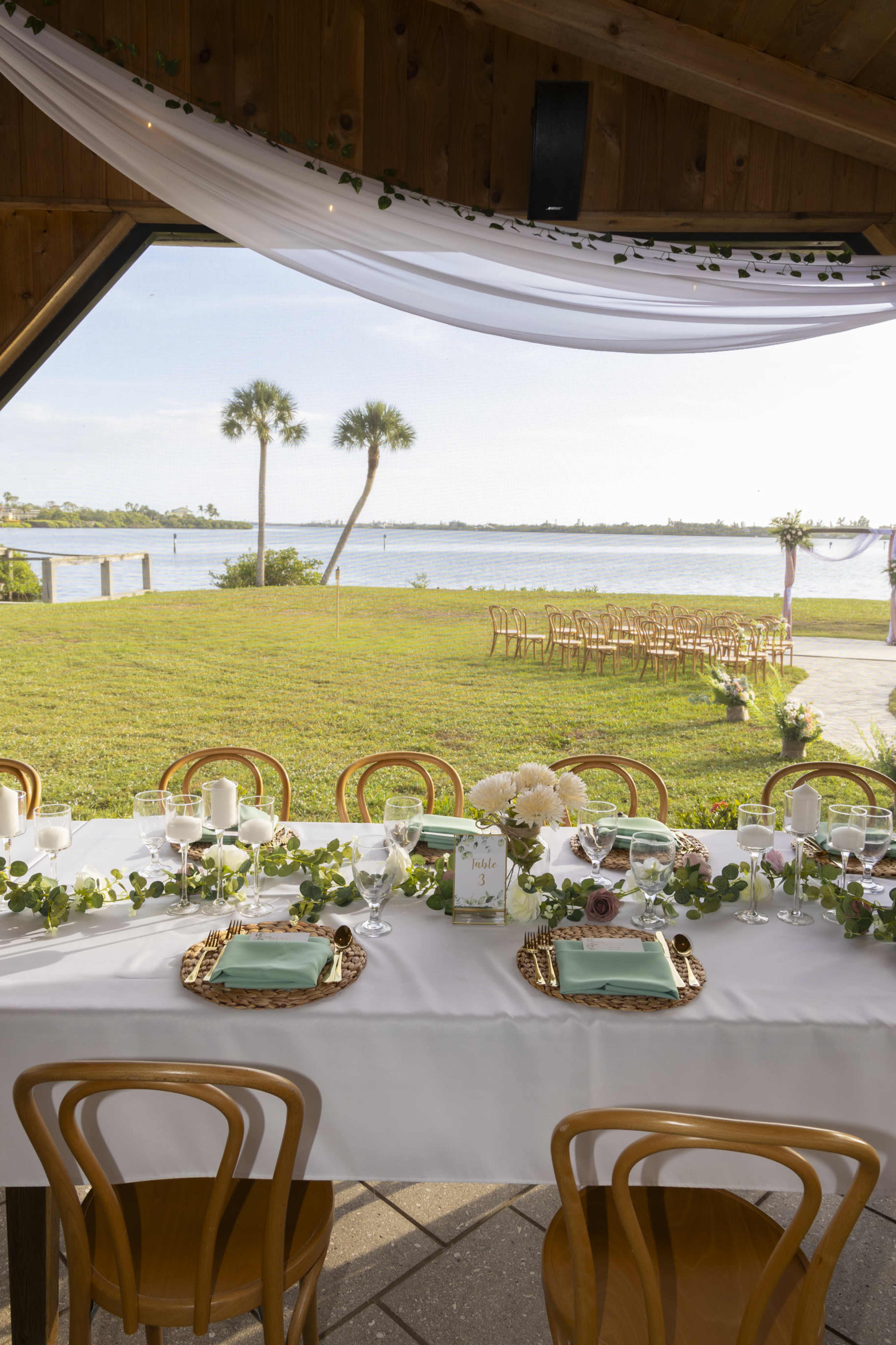 A decorated table is set for a gathering next to a water view, surrounded by greenery and chairs arranged for guests.