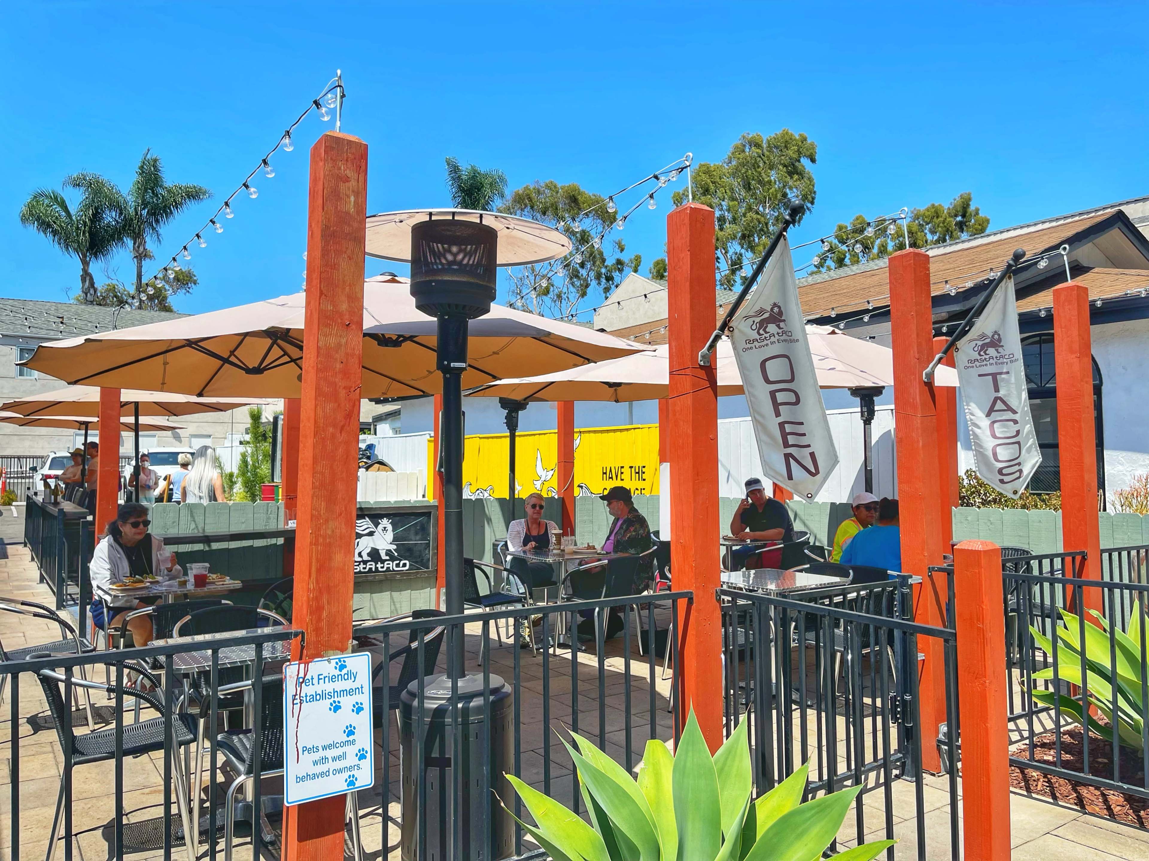 A bustling outdoor patio at a restaurant features diners seated under umbrellas, with flags displaying "Open" and "Tacos" prominently displayed.