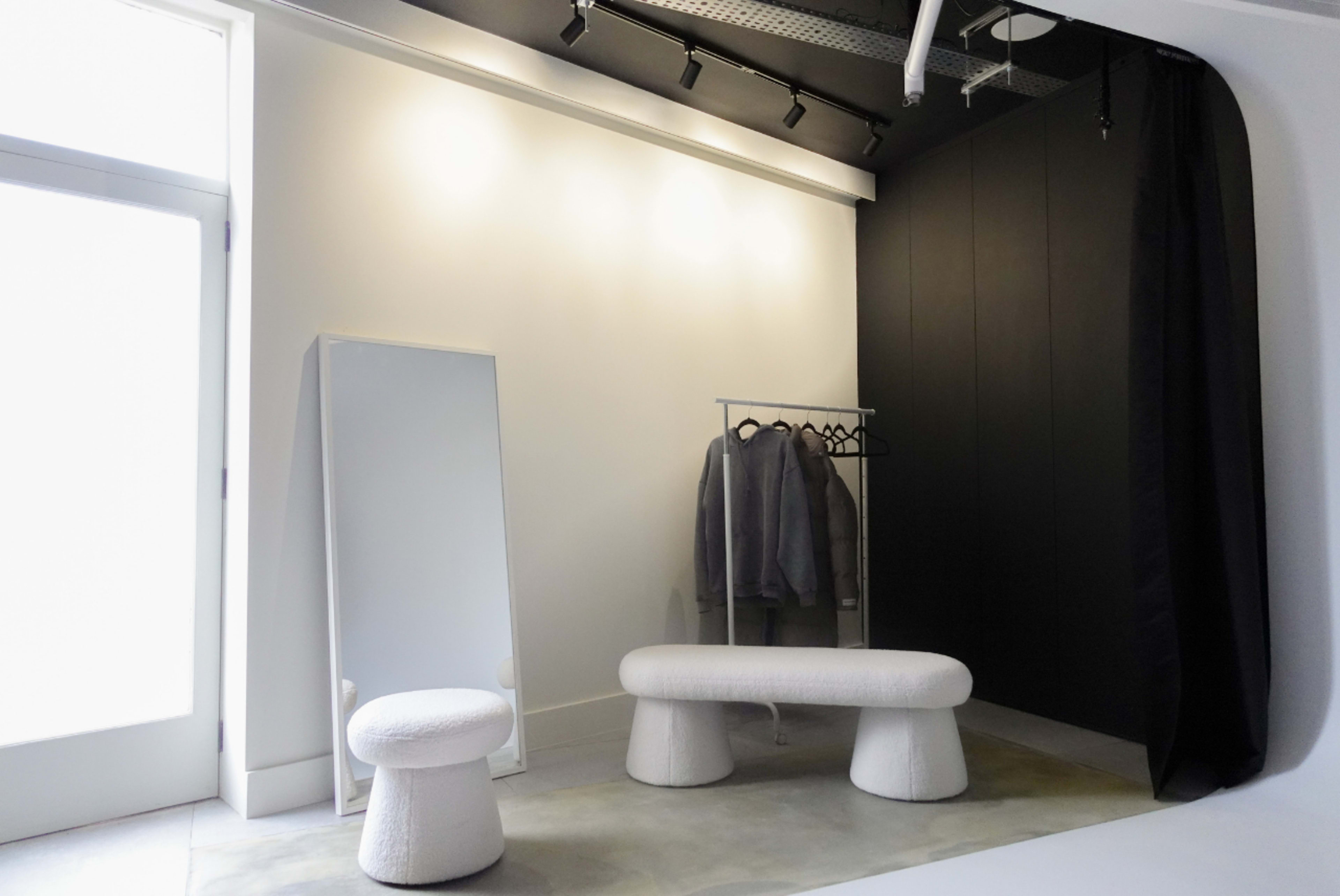 A minimalist dressing area featuring a full-length mirror, a clothing rack with gray garments, and two white, sculptural seating pieces.