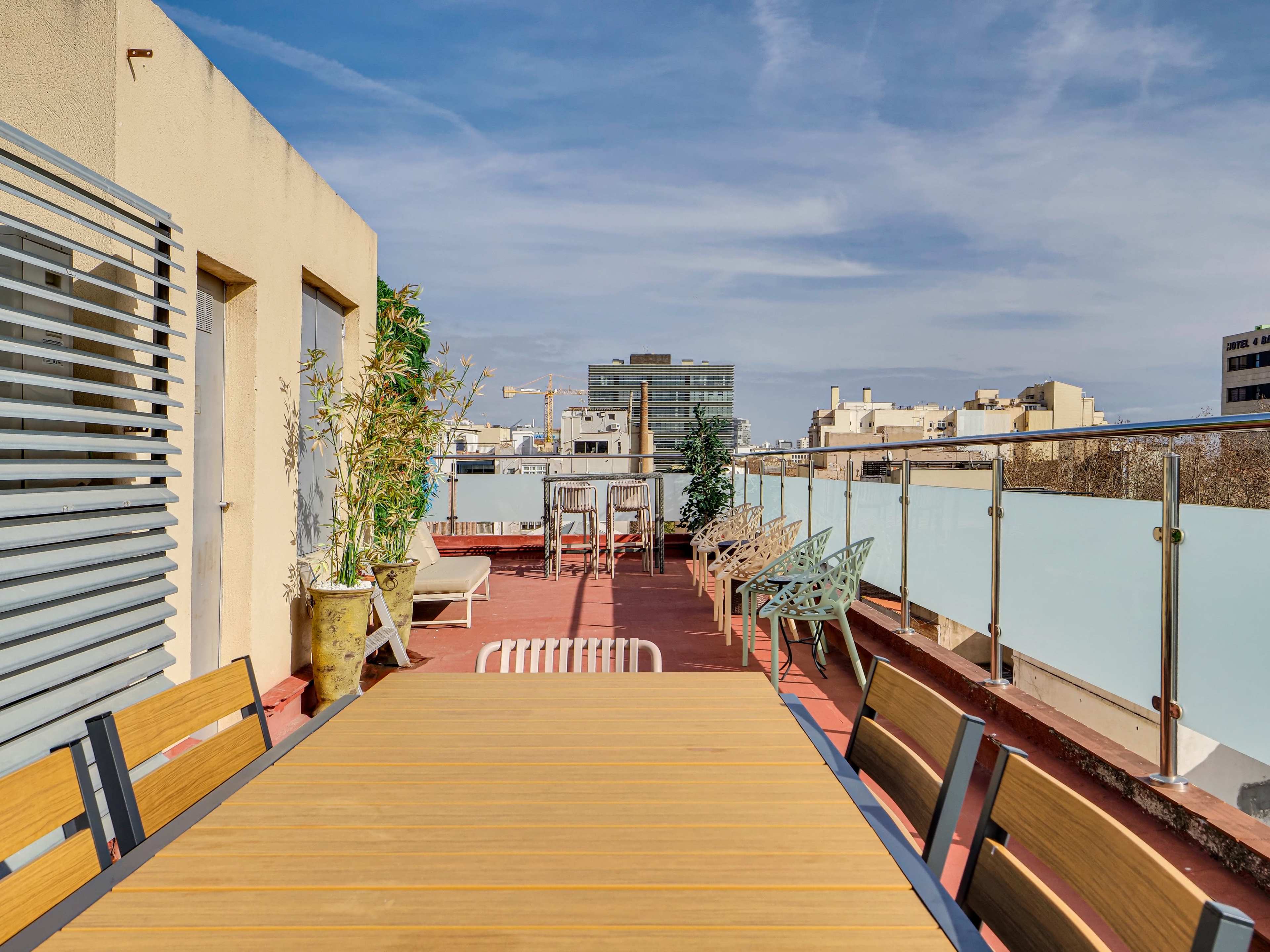 A spacious rooftop terrace features a long wooden table surrounded by chairs, with city buildings and a blue sky in the background.