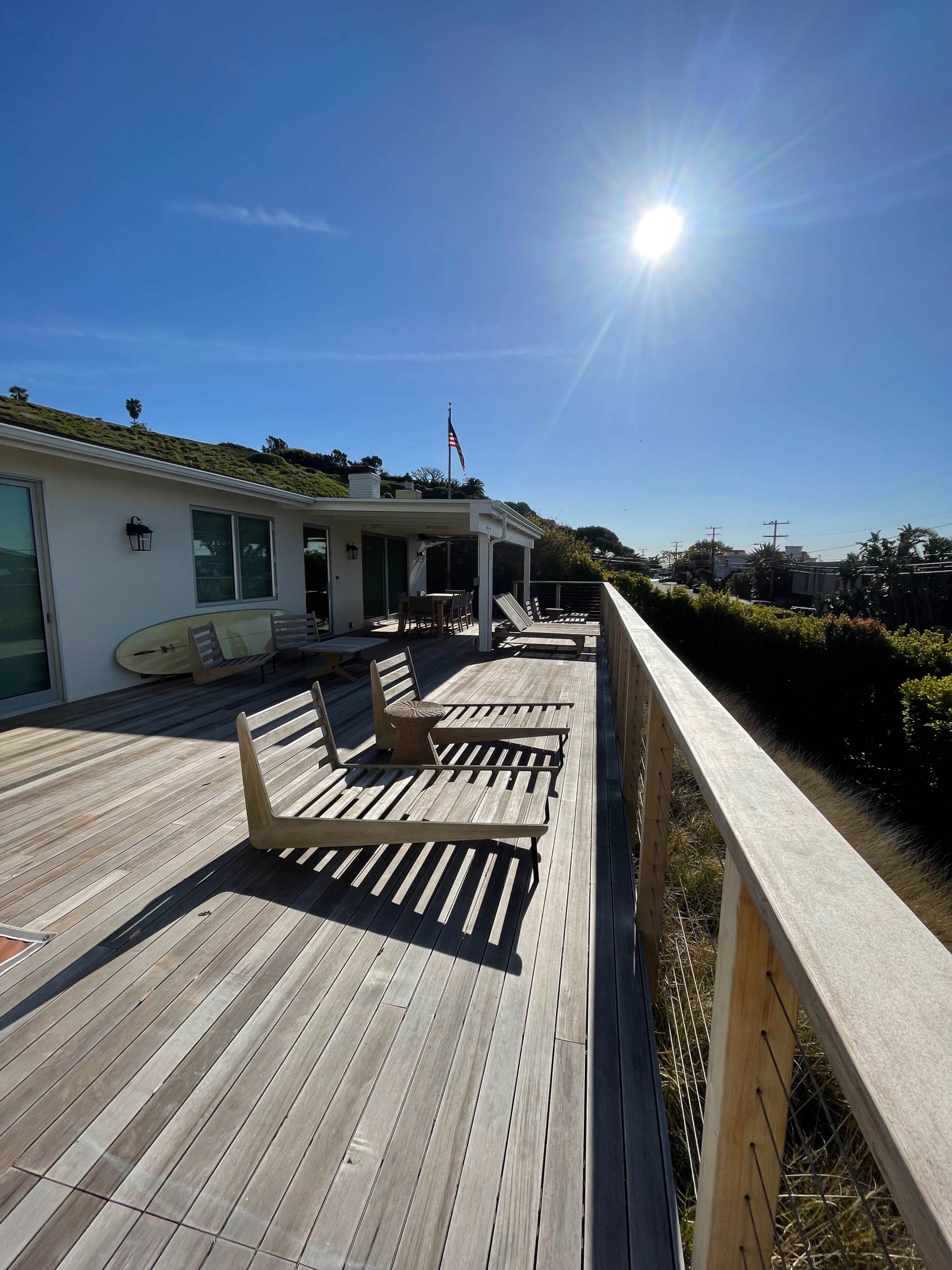 A spacious wooden deck with lounge chairs, a surfboard propped against the wall, and a clear blue sky with the sun shining brightly.