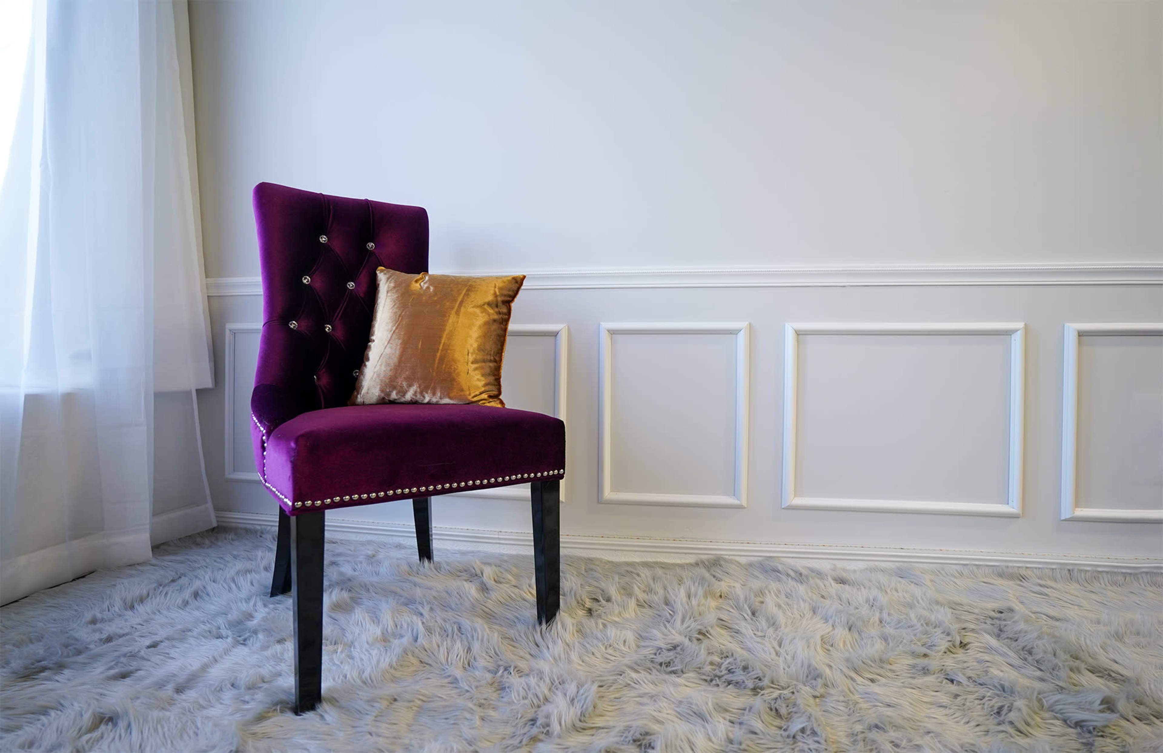 A purple upholstered chair with a gold decorative pillow sits on a plush rug in a softly lit room with white paneling.