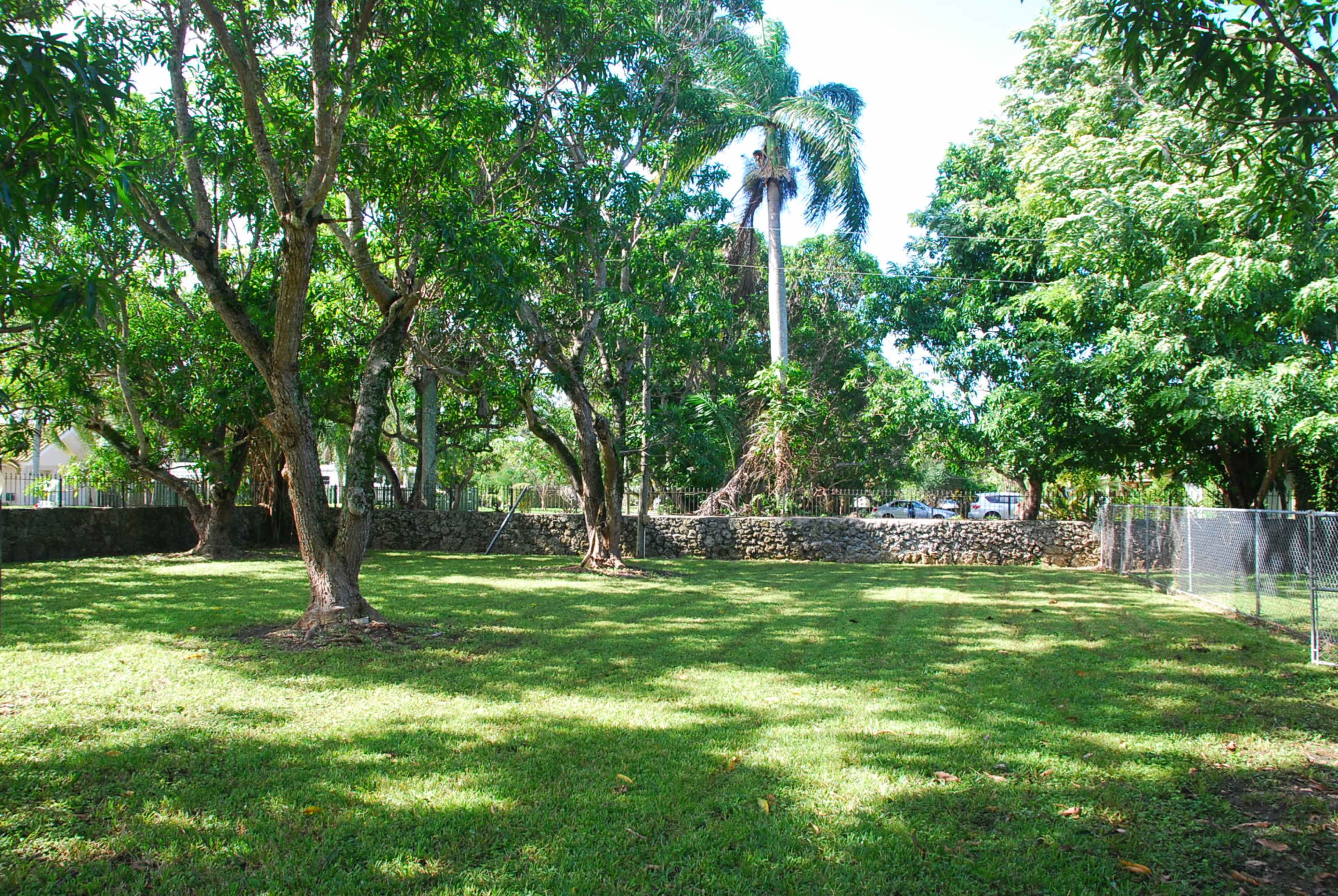 A grassy area surrounded by trees and a stone wall, with a fence along one side.