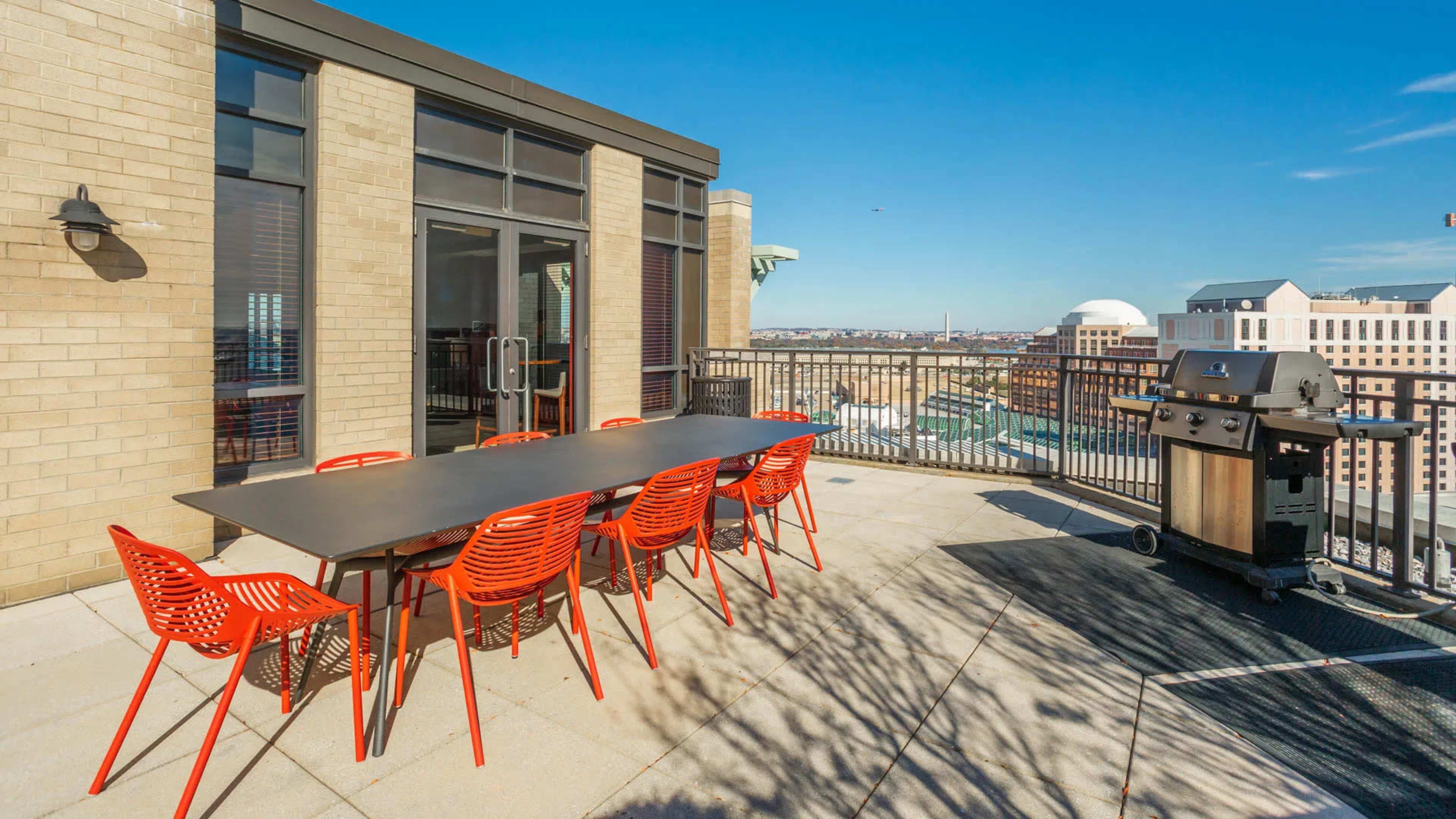 A rooftop patio features a large table with red chairs and a grill, overlooking the city skyline.