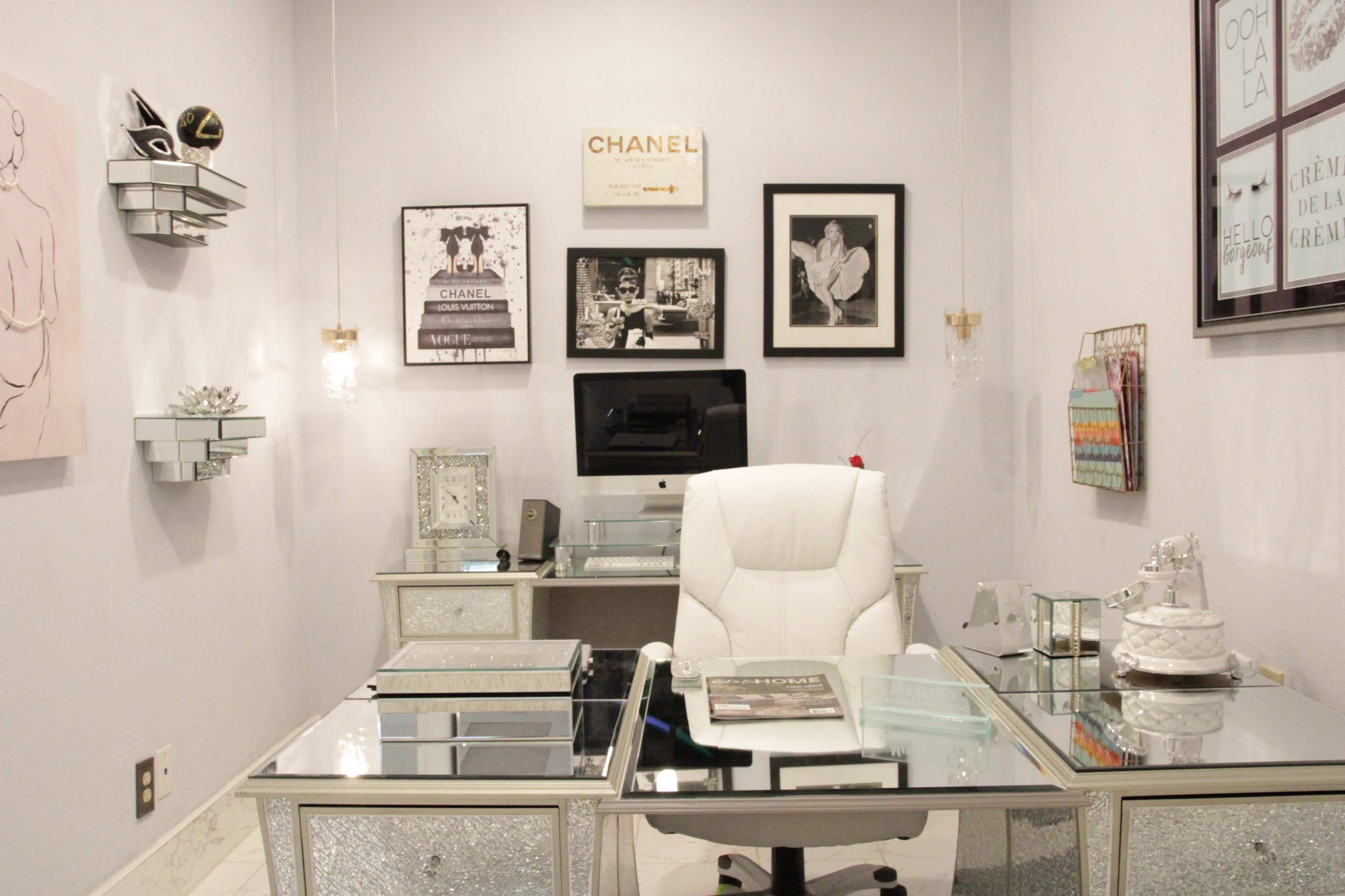 A modern office with a mirrored desk, an ergonomic white chair, and framed artwork on the walls, including a Chanel print.