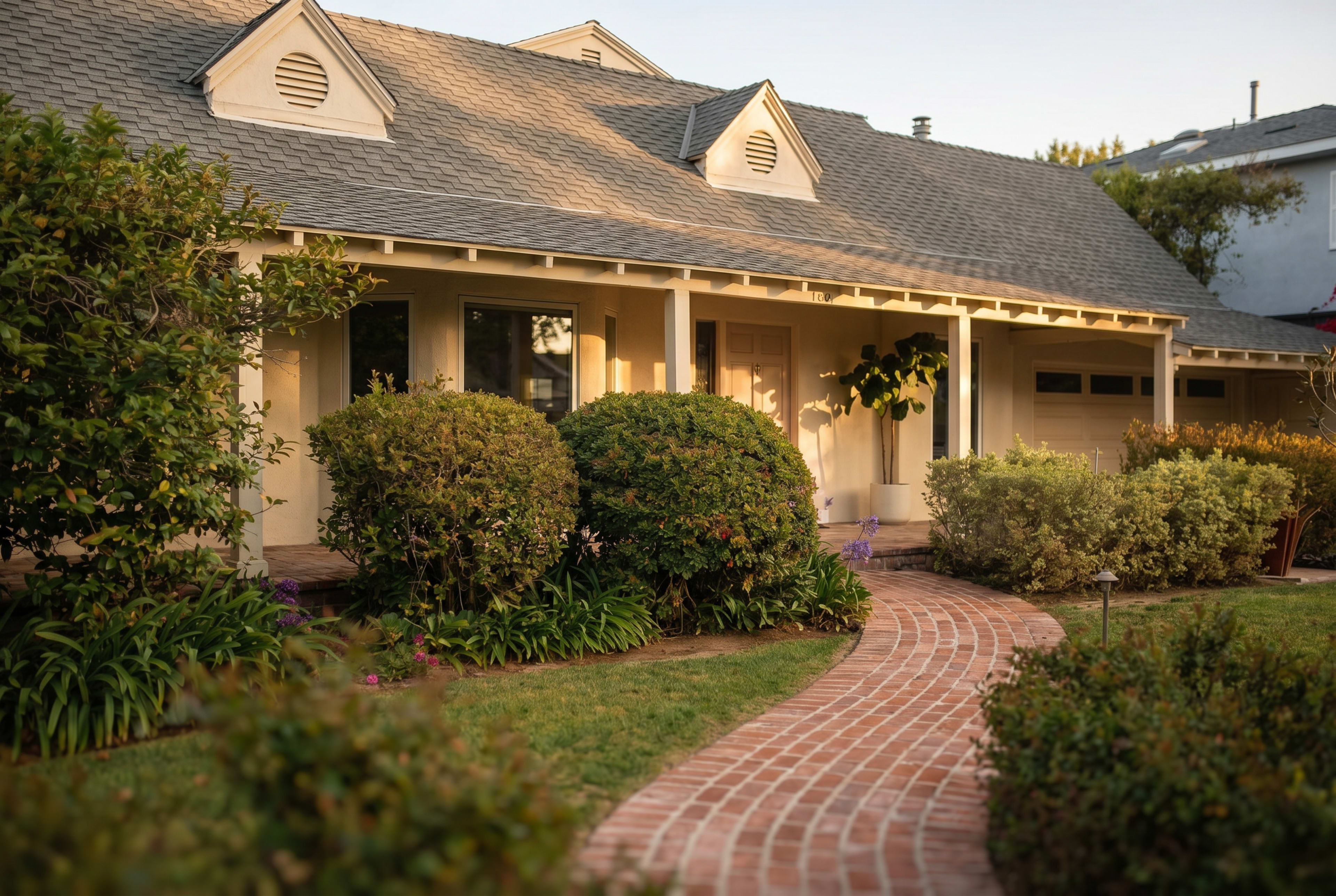 The image depicts a residential home with a brick walkway leading to a covered porch, surrounded by well-maintained shrubs and plants.