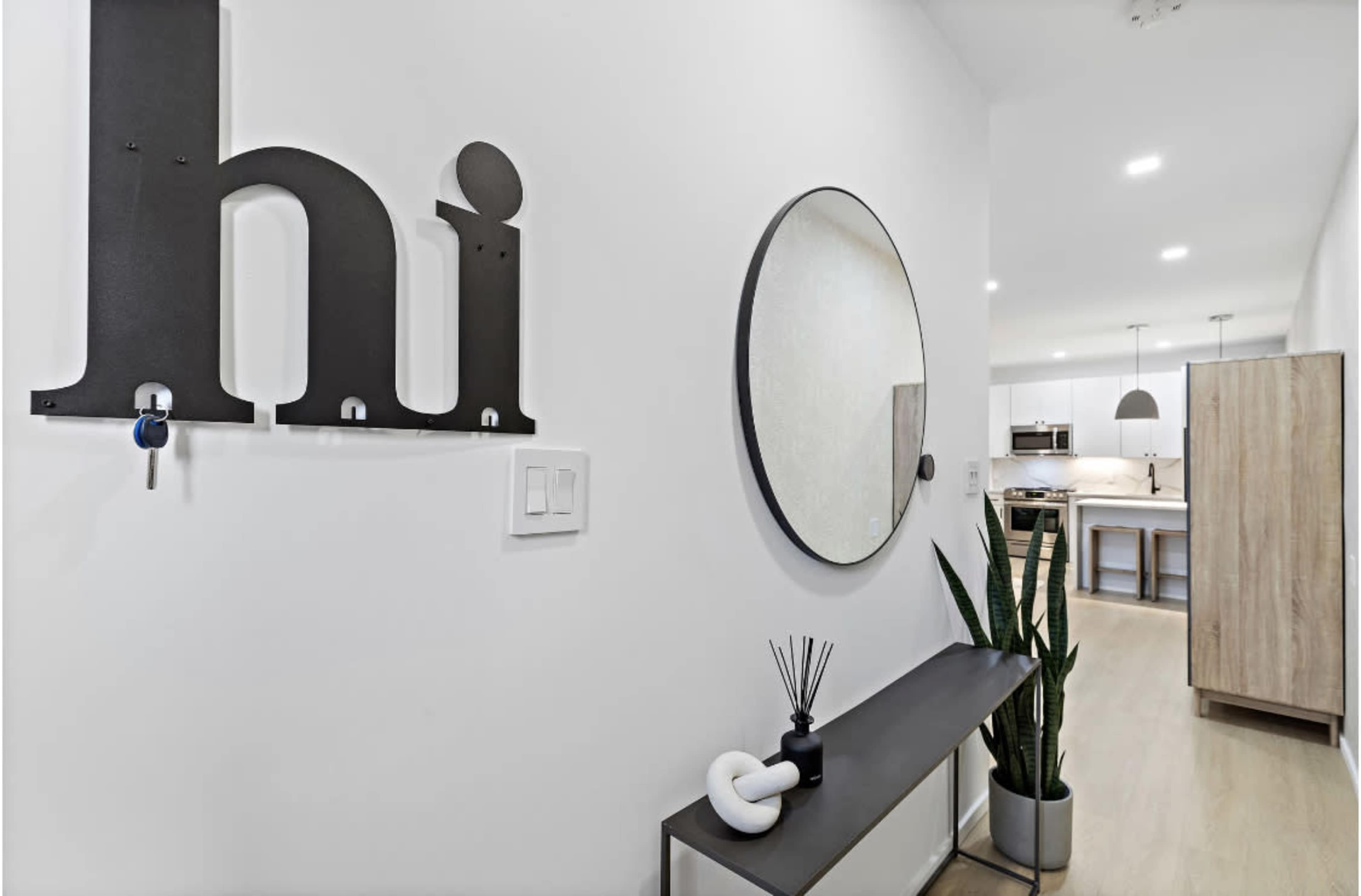 An entryway with a wall-mounted "hi" sign, a round mirror, a wooden console table with a plant, and a view of a kitchen in the background.