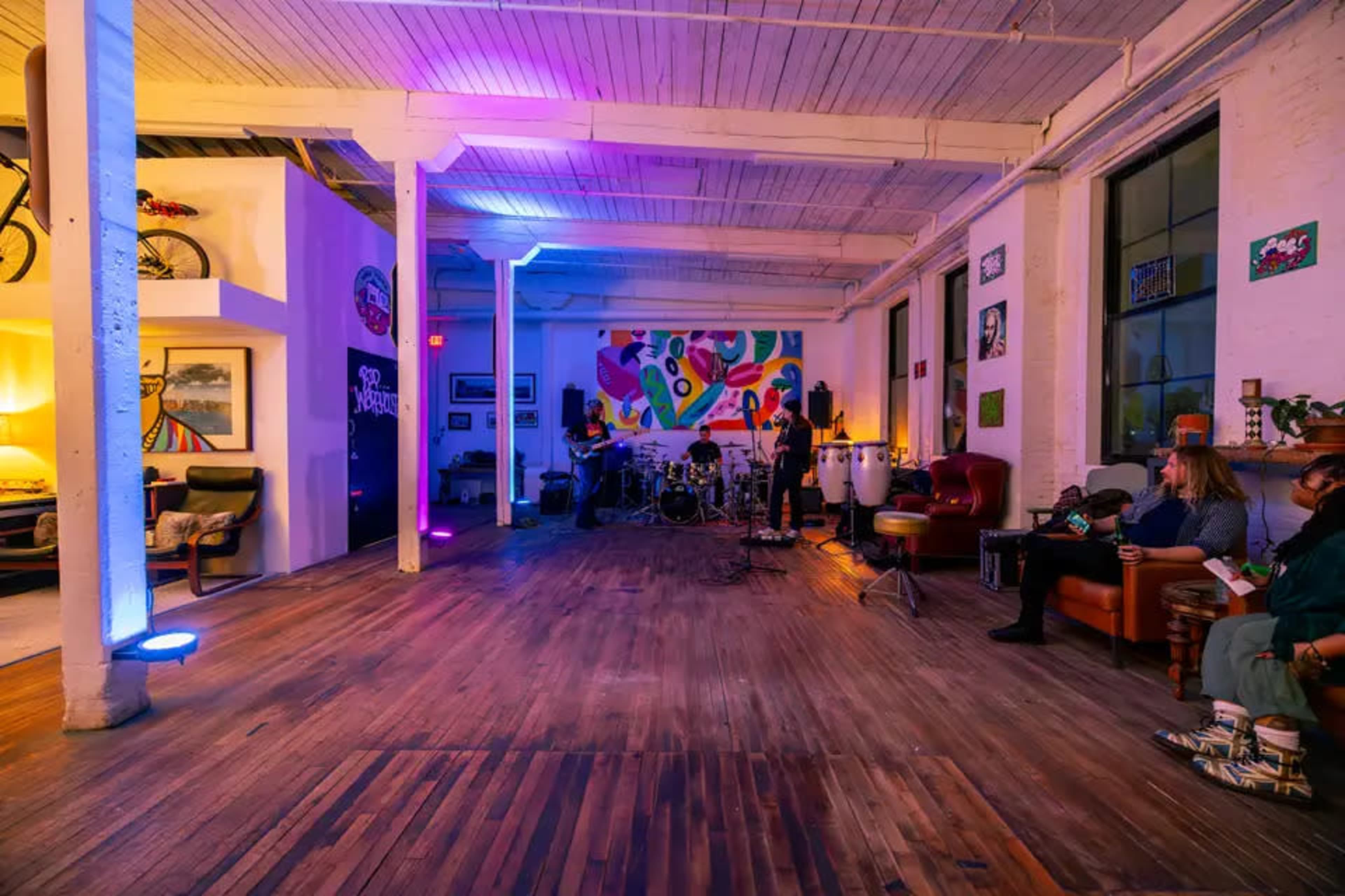 The image shows a spacious room with wooden flooring, featuring musical instruments set up in the center and colorful lighting illuminating the walls.