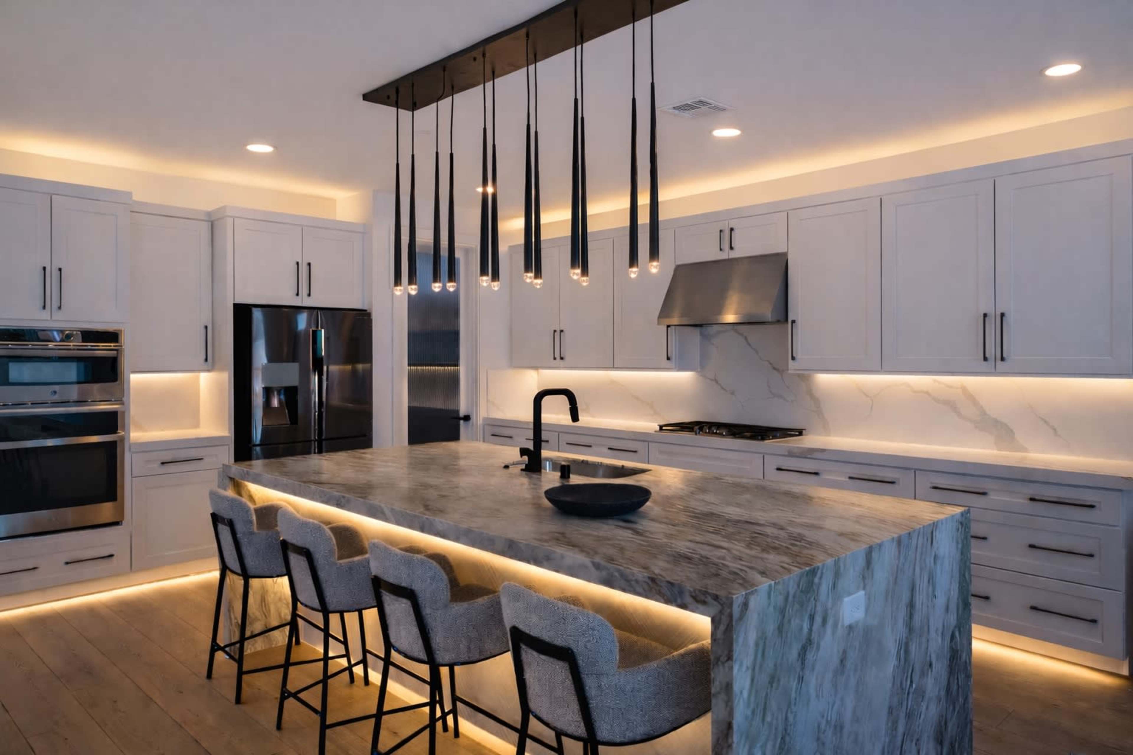 A modern kitchen features a large marble island with bar seating, surrounded by sleek cabinetry and illuminated by pendant lights.