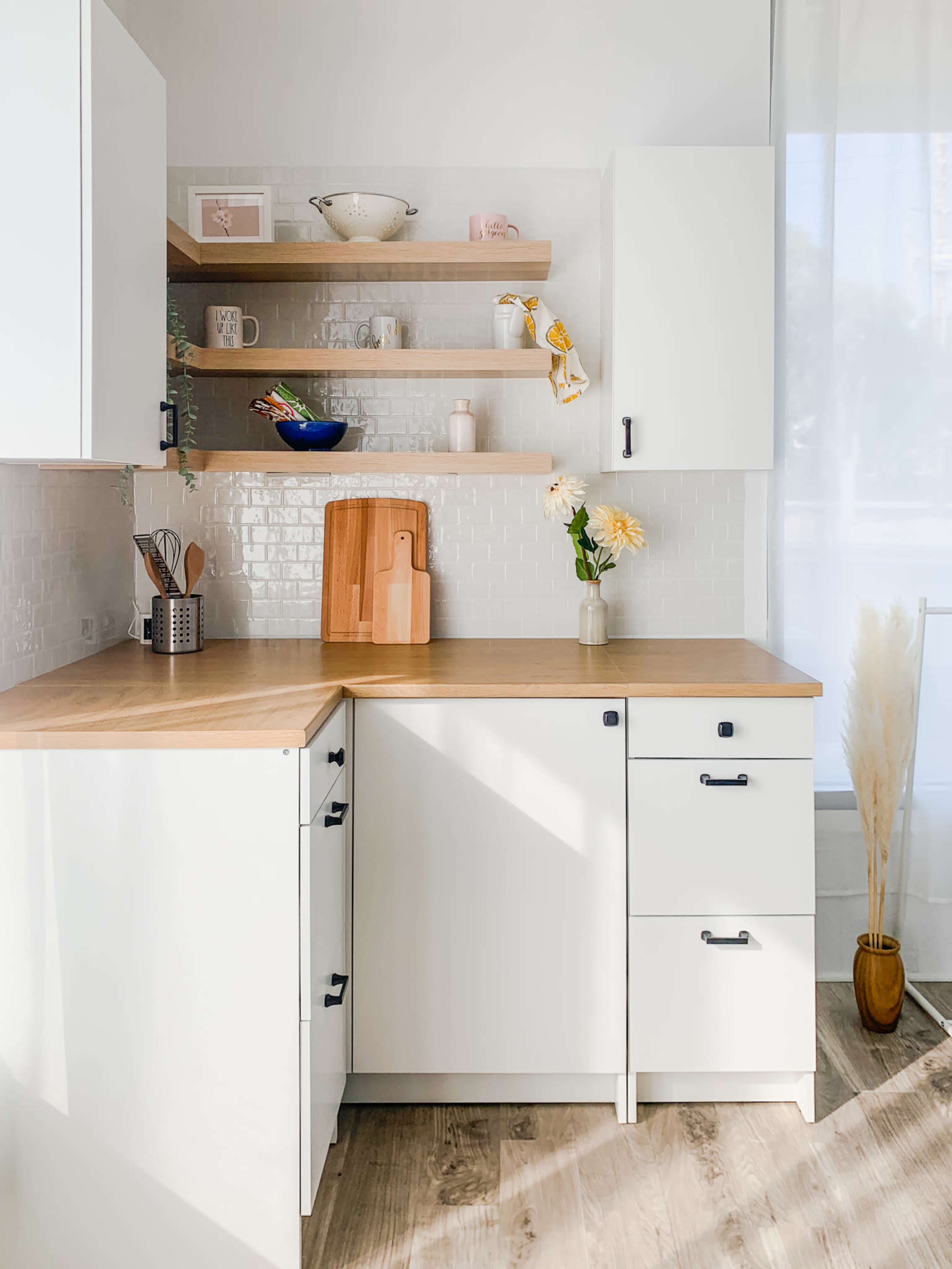 A bright kitchen features white cabinetry, wooden shelves, and a wooden countertop, with decorative items and a vase of flowers on display.