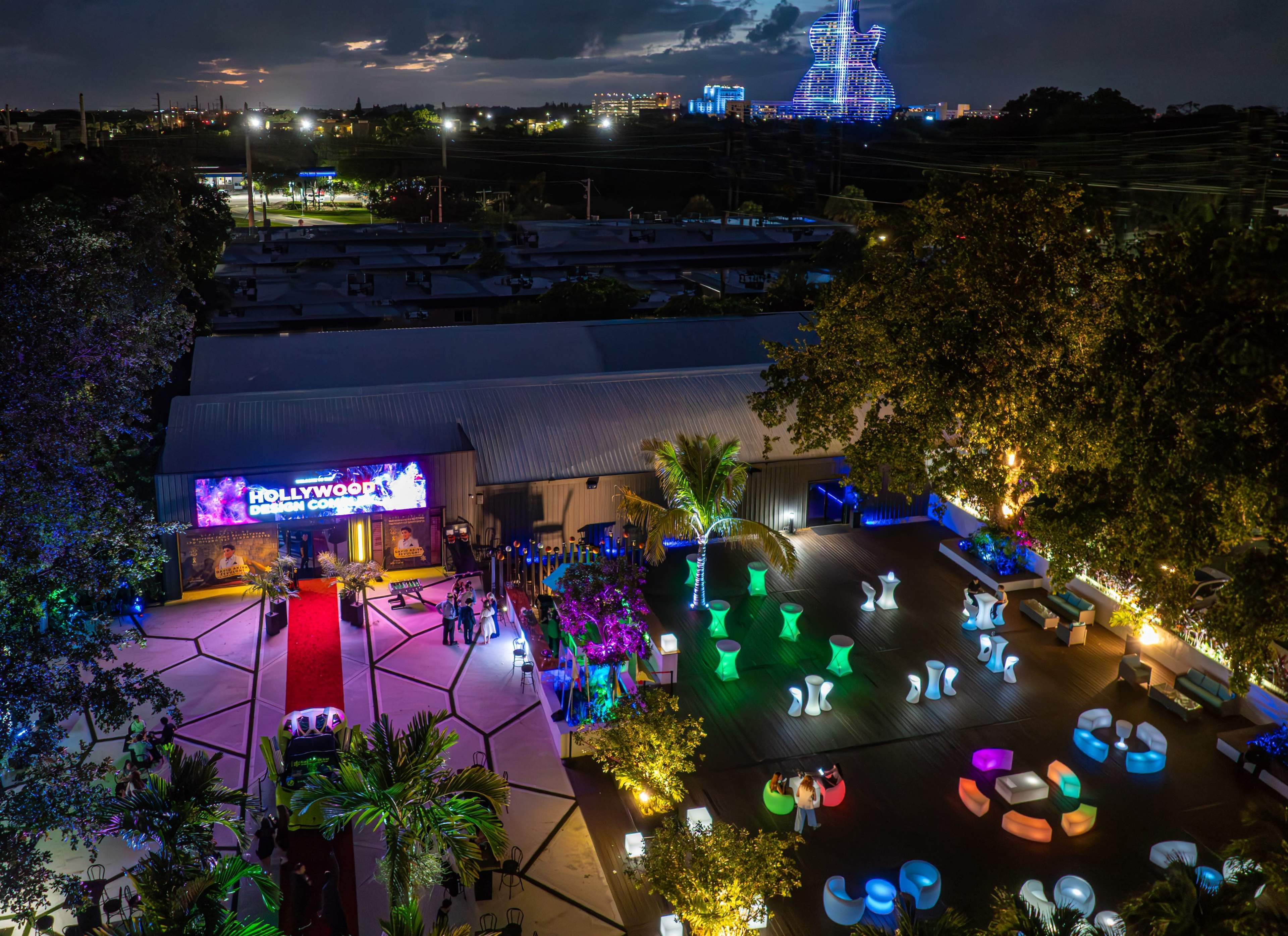 The image shows a brightly lit outdoor event space at night, featuring colorful seating arrangements and a stage with a sign that reads "HOLLYWOOD" in front of a crowd of palm trees.