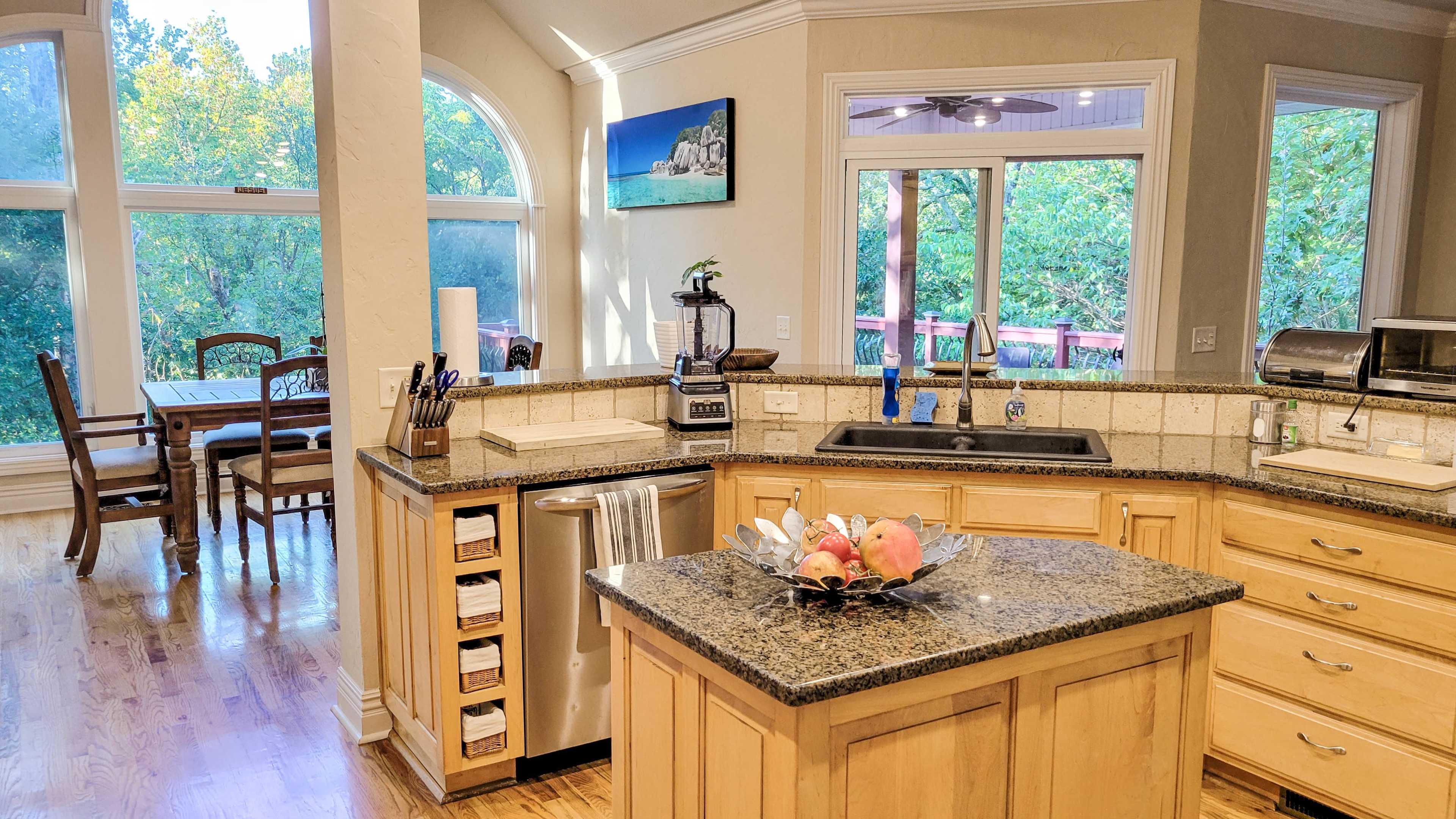 A modern kitchen with wooden cabinetry, a granite countertop, and a dining area visible through large windows.