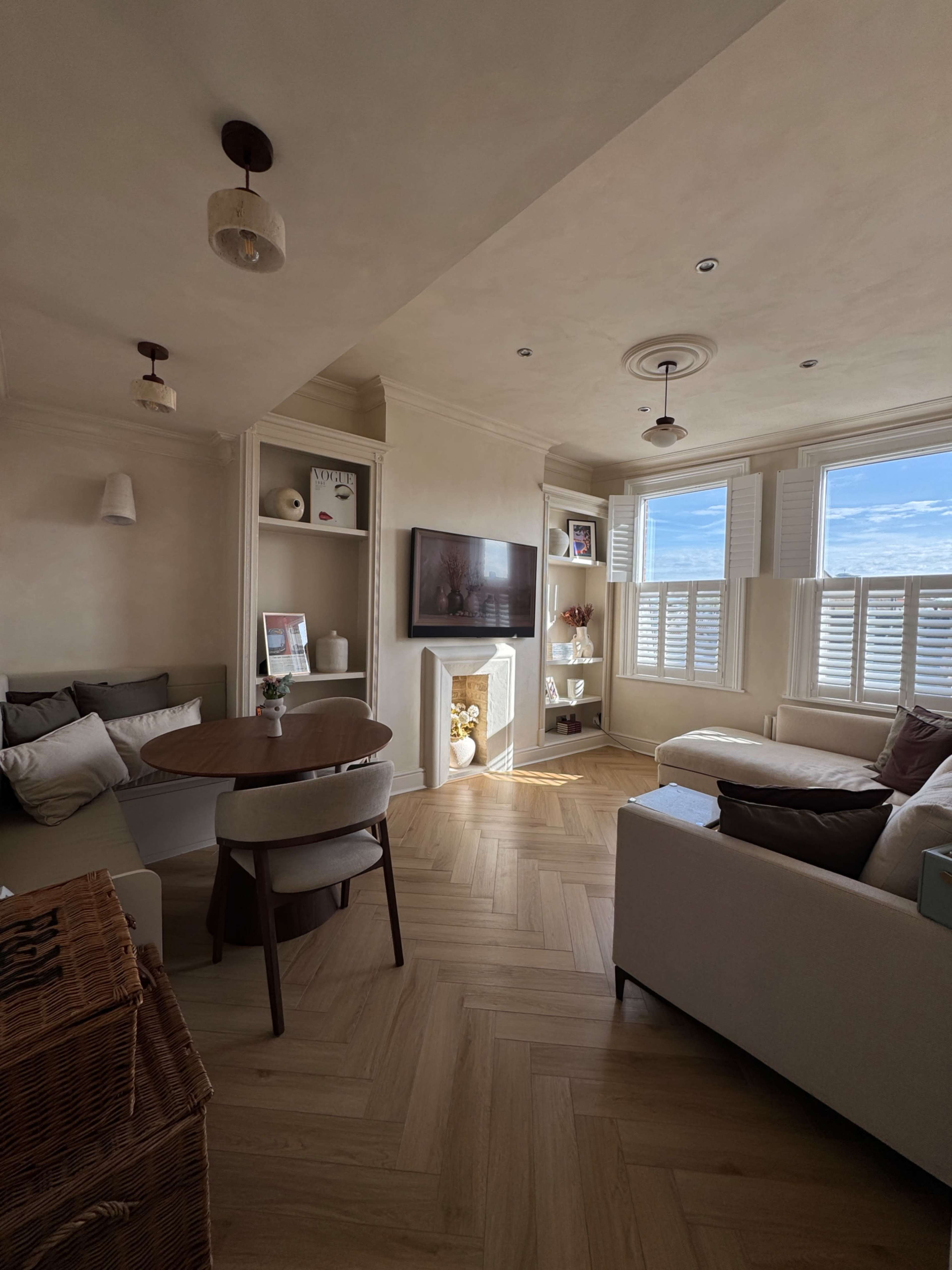 The image shows a cozy living room with a circular dining table, a white sofa, and shelves displaying decor, illuminated by natural light through window shutters.