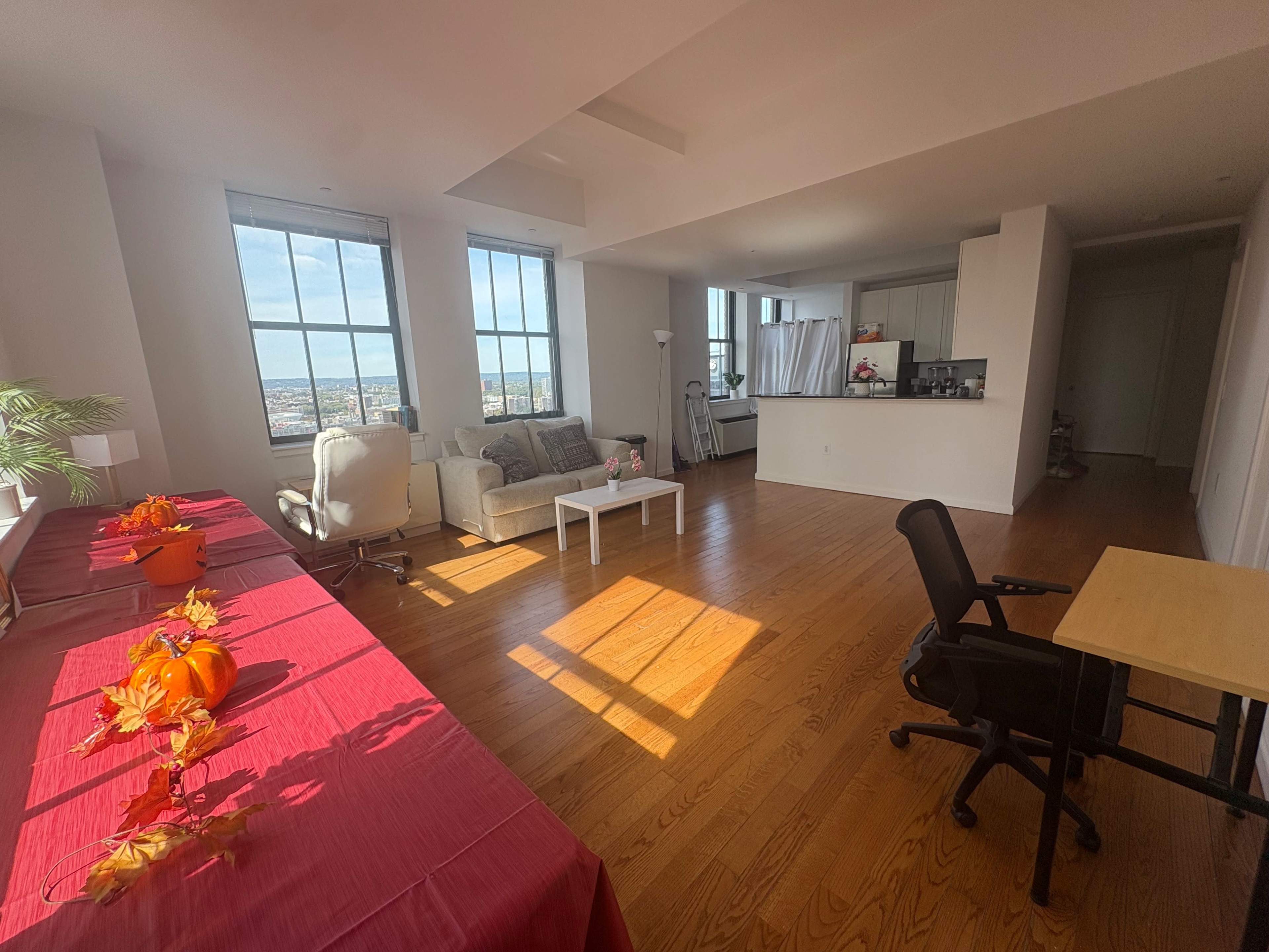 The image shows a bright, spacious living area with hardwood floors, large windows, a beige couch, a small dining table with decorative items, and a work desk.