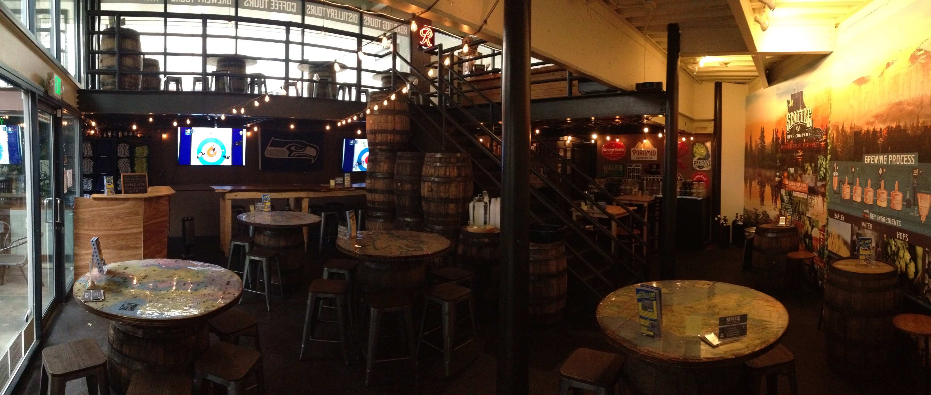 A bar interior with wooden barrel tables, upstairs seating, and screens showing sports events.