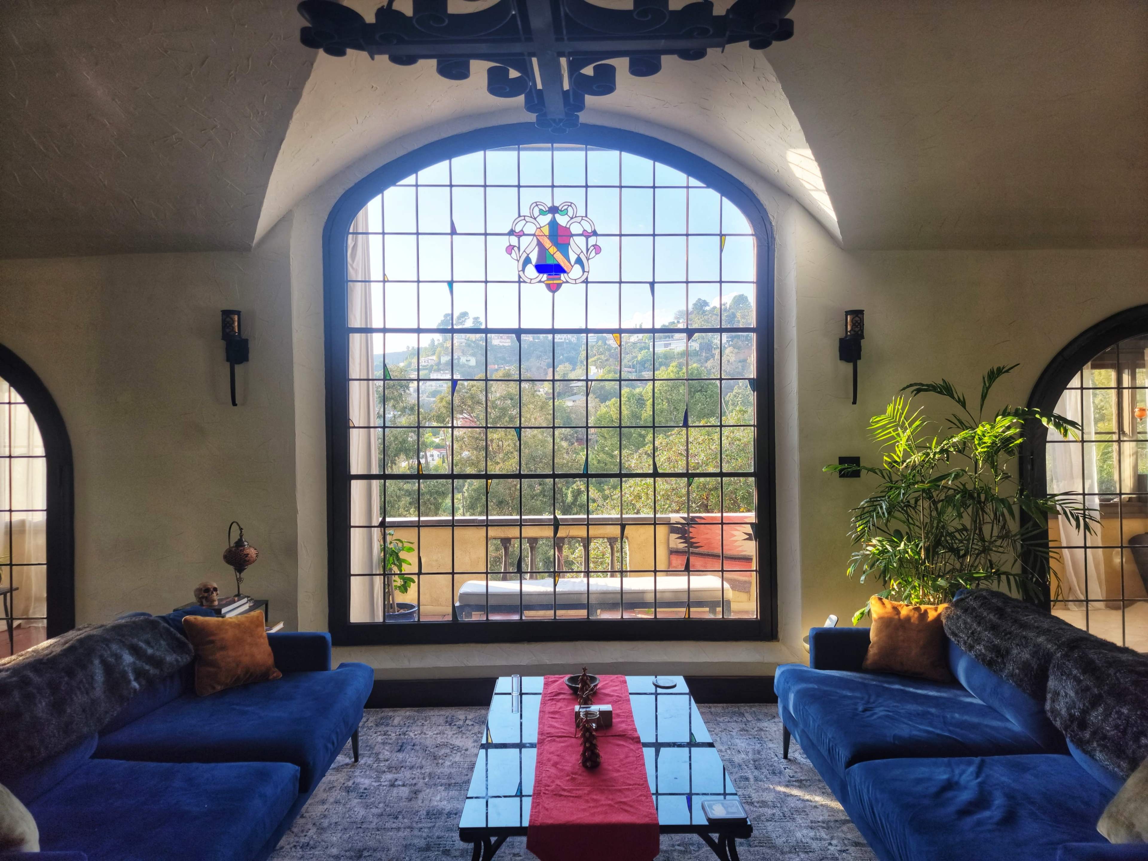 A living room features blue sofas and a coffee table in front of a large arched window with a stained glass design, overlooking a scenic view.