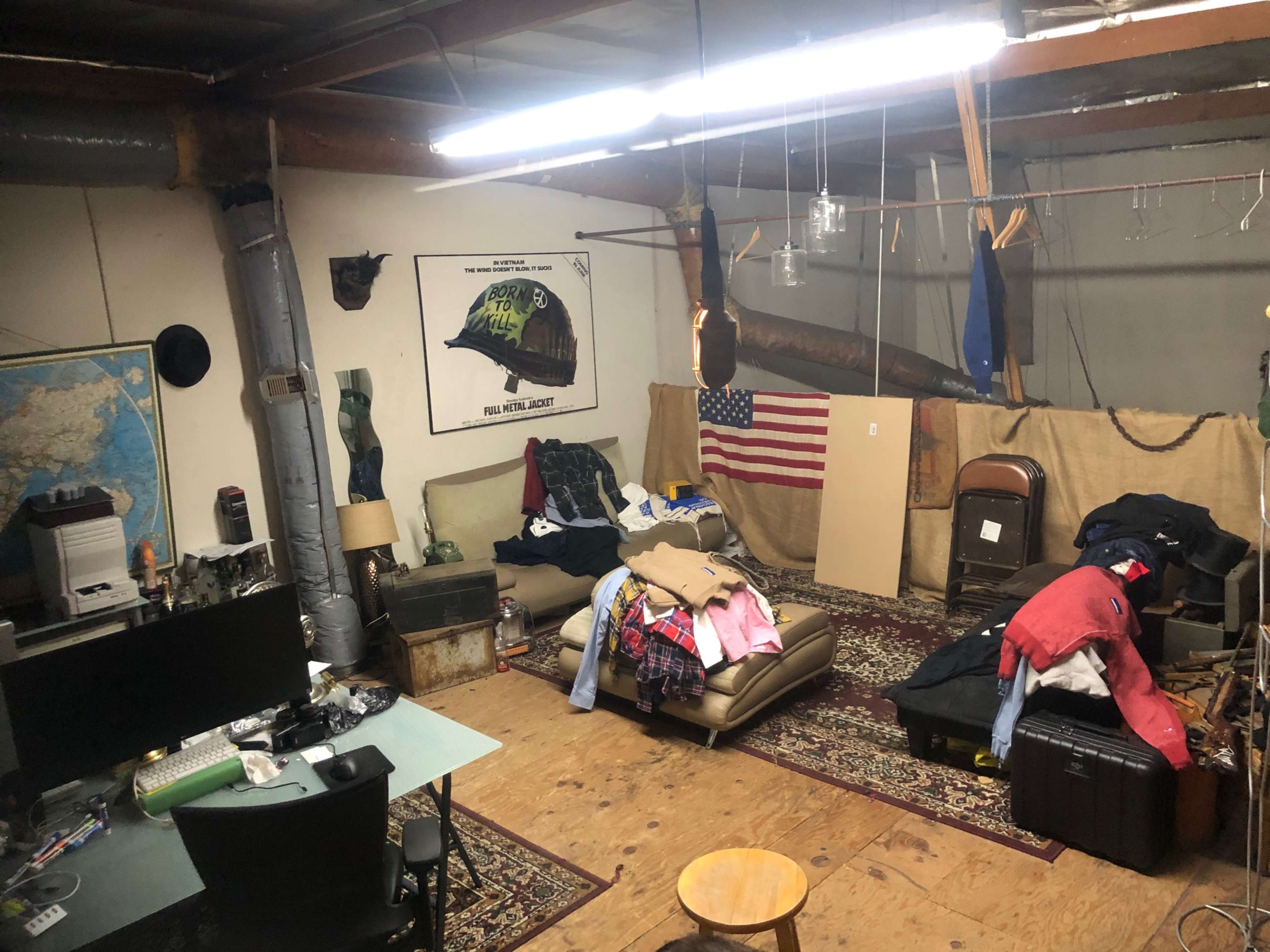 A cluttered room with a sofa, scattered clothing, an American flag, a desk with a computer, and various items on the floor and walls.