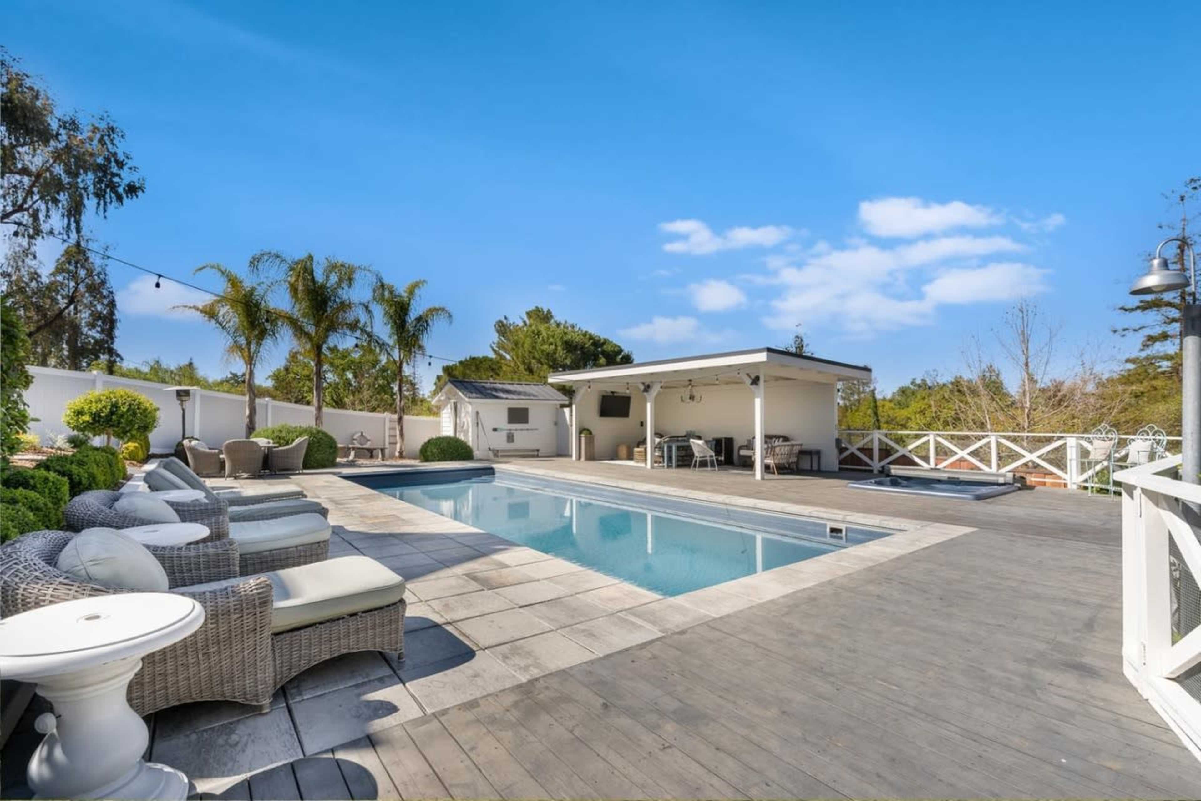 The image shows a backyard with a swimming pool surrounded by wooden decking, lounge chairs, and a poolside cabana.