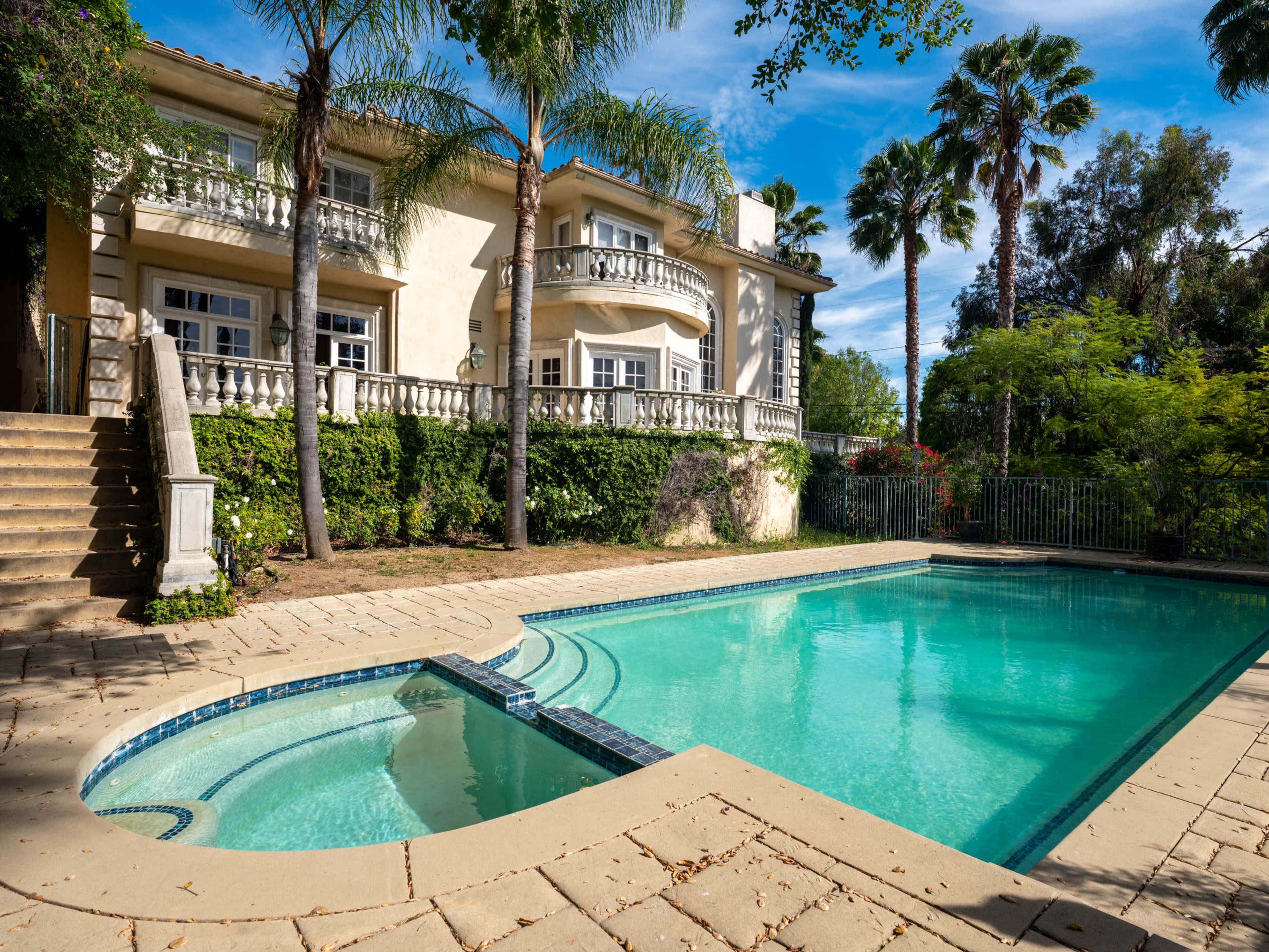 The image shows a large house with a swimming pool in the foreground, surrounded by palm trees and landscaped greenery.