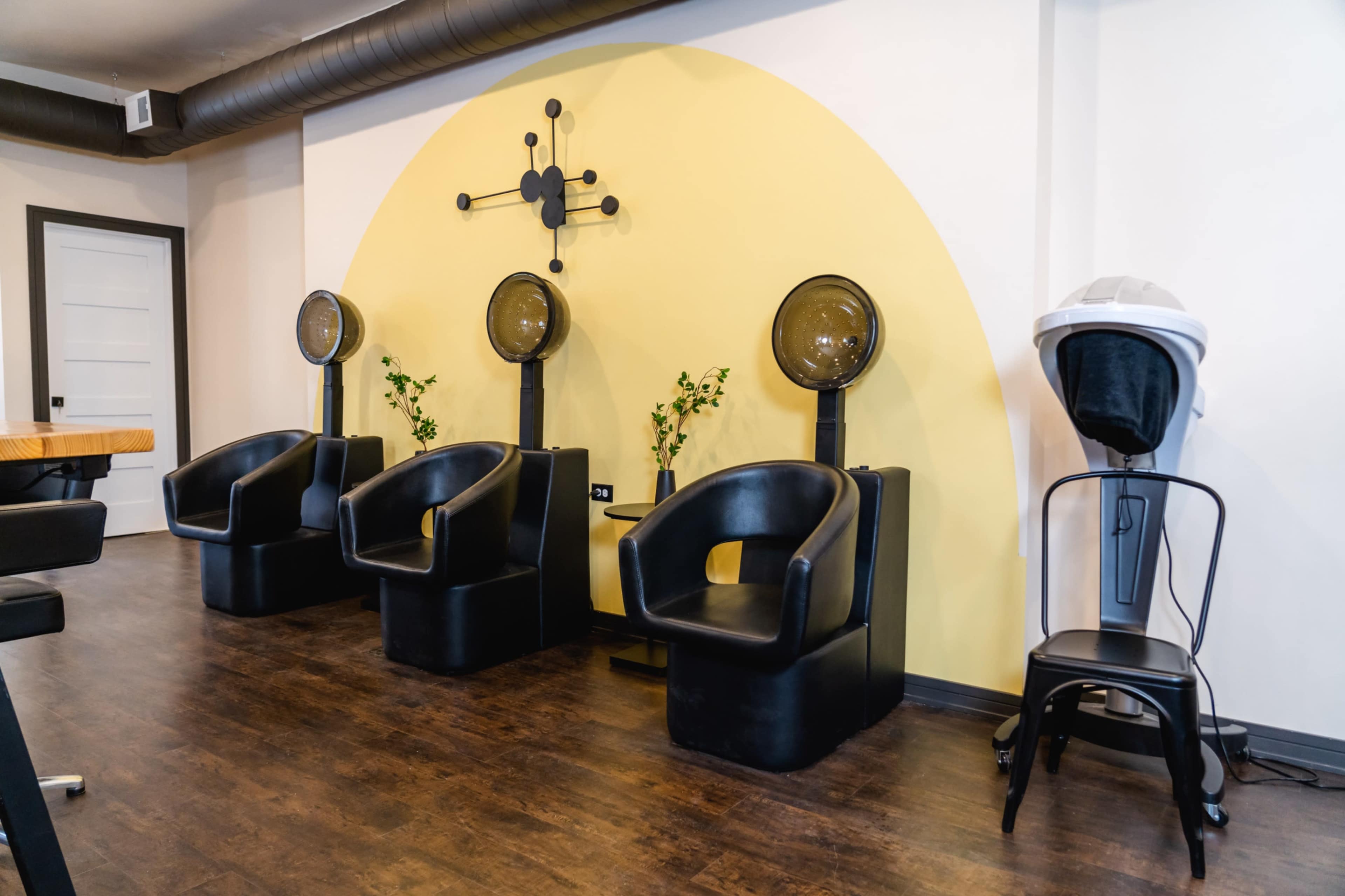 A salon interior featuring three black salon chairs with hair dryers positioned against a yellow semicircular wall.
