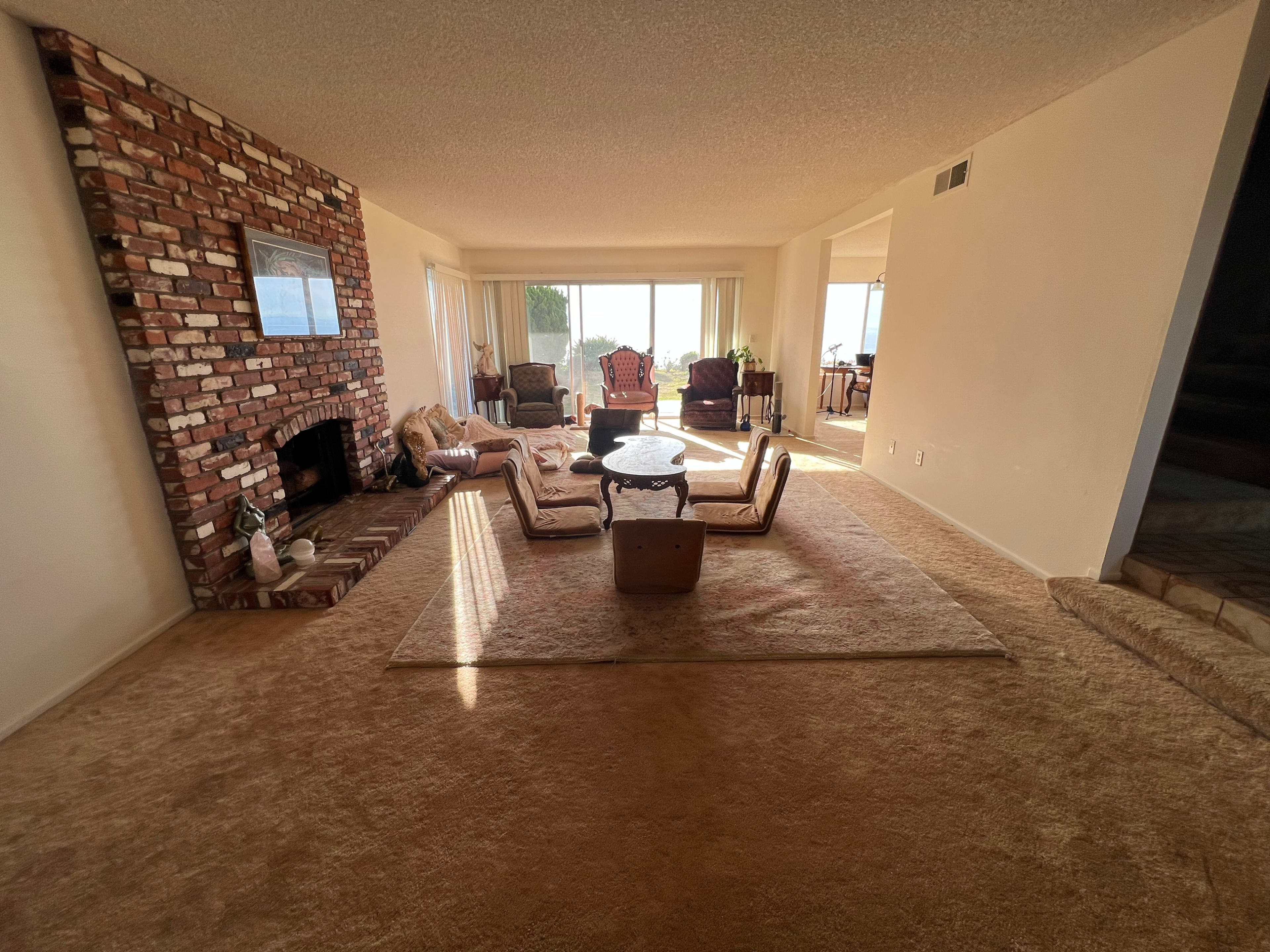 The image depicts a spacious living room featuring a brick fireplace, a large area rug, and several pieces of furniture arranged around a central coffee table, with large windows allowing natural light to fill the space.